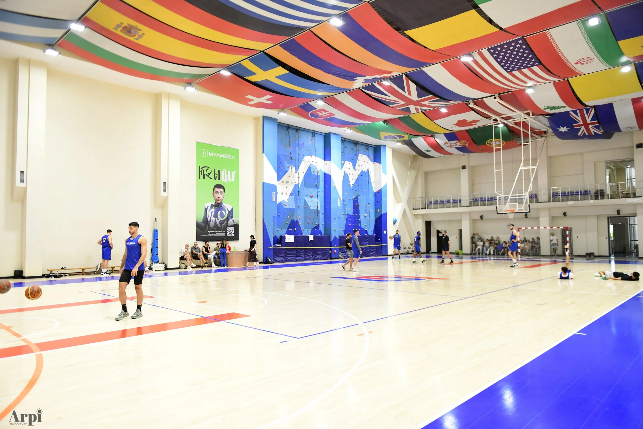 Indoor sports gym with international flags on the ceiling, people playing basketball, and a climbing wall visible on the side wall.