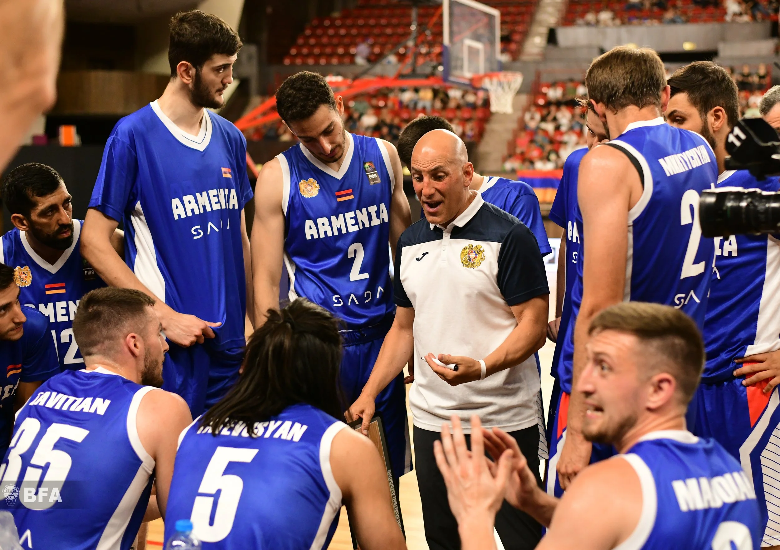 Basketball coach giving a team talk to players wearing "Armenia" jerseys during a game in an indoor stadium.