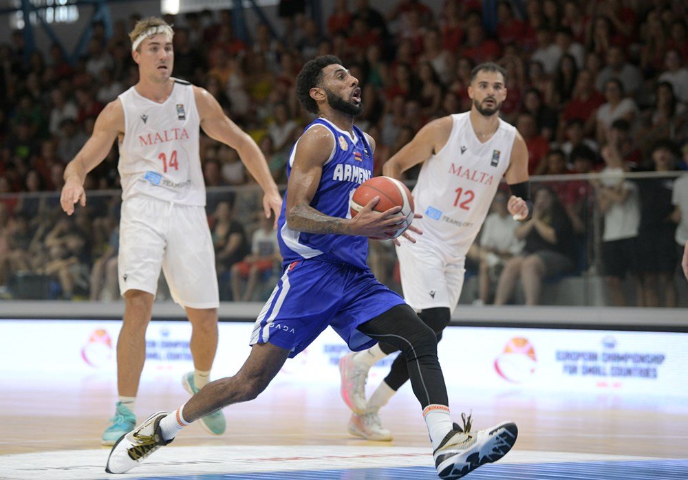 Basketball player in blue jersey from Armenia holding the ball while running during a game against Malta players dressed in white jerseys, in a crowded indoor stadium.