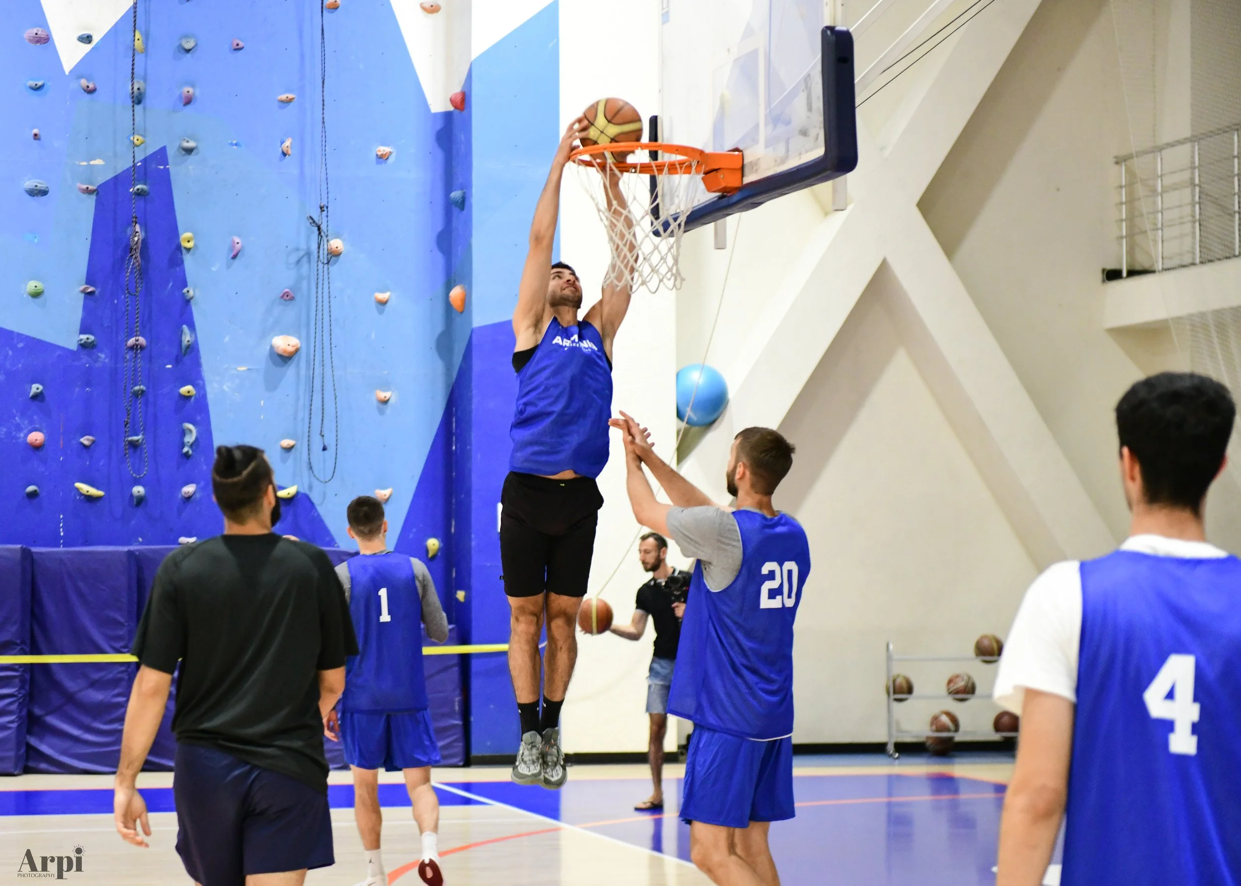 Basketball players practicing in a gym with a climbing wall in the background. One player is mid-air making a dunk, while others watch. They're wearing blue jerseys with numbers.