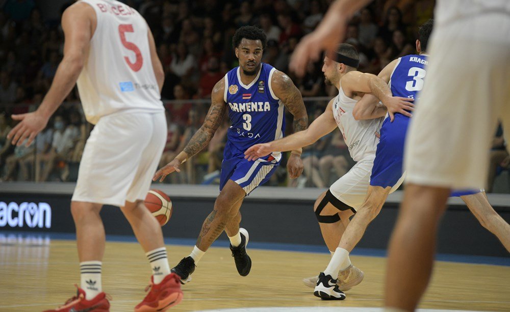 Basketball game scene featuring players from Armenia in blue jerseys and another team in white jerseys during a match, with one player dribbling the ball.