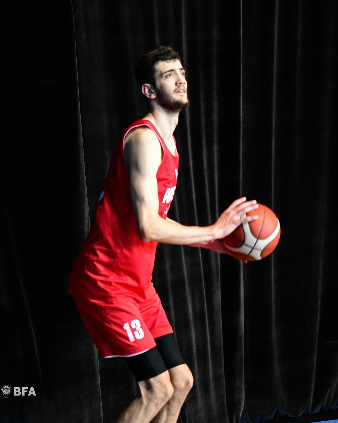 Basketball player preparing to shoot, wearing red uniform with number 13 against a dark curtain background.