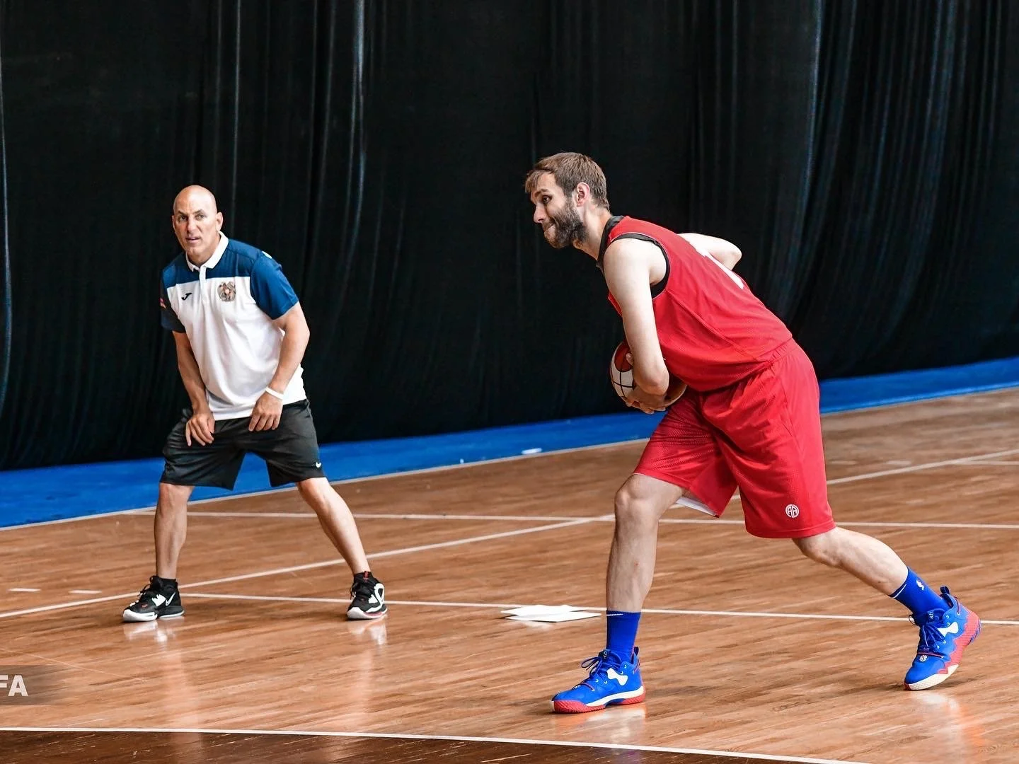 Two men on a basketball court; one wearing a red uniform holding a basketball, the other in a white and blue shirt and shorts coaching or watching.