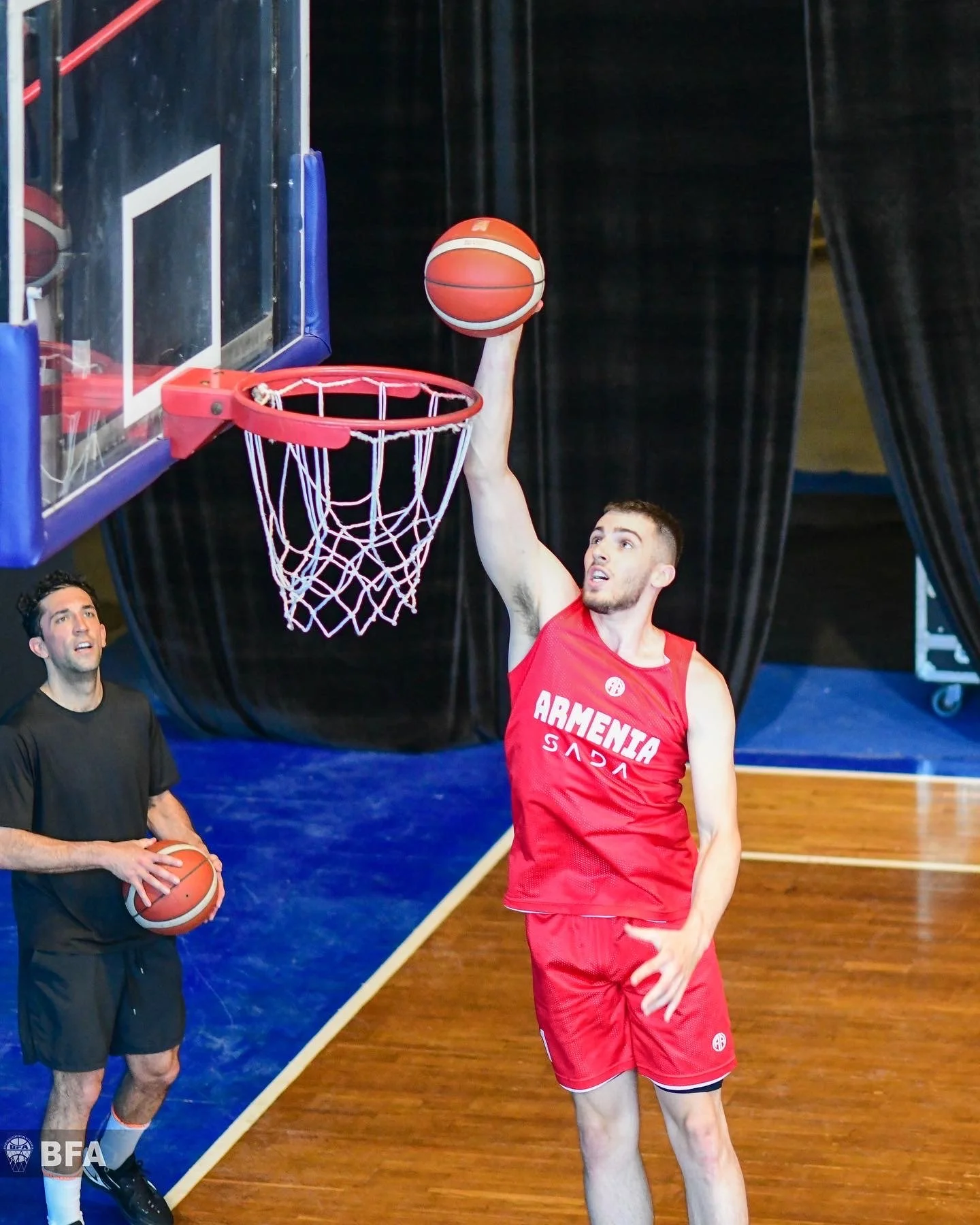 Two basketball players on an indoor court. One player in red jersey performs a layup, while another, in black, holds a basketball and watches.