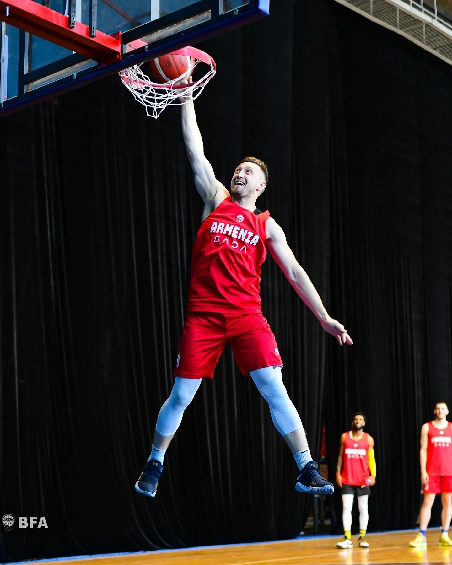 Basketball player wearing a red "Armenia" jersey dunking a ball in an indoor court.