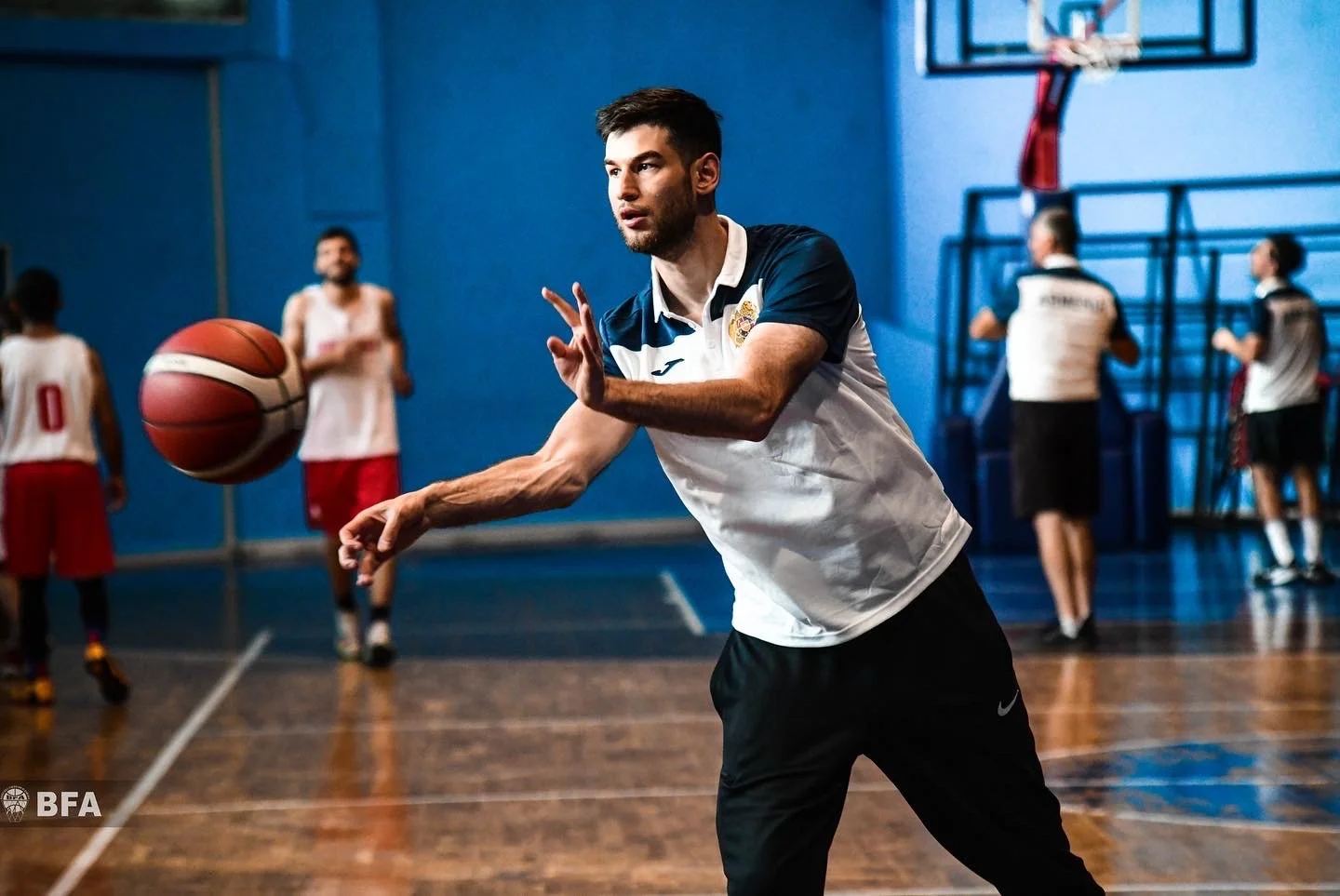 A man in a sports jersey and shorts passing a basketball in an indoor court with several other players in the background.