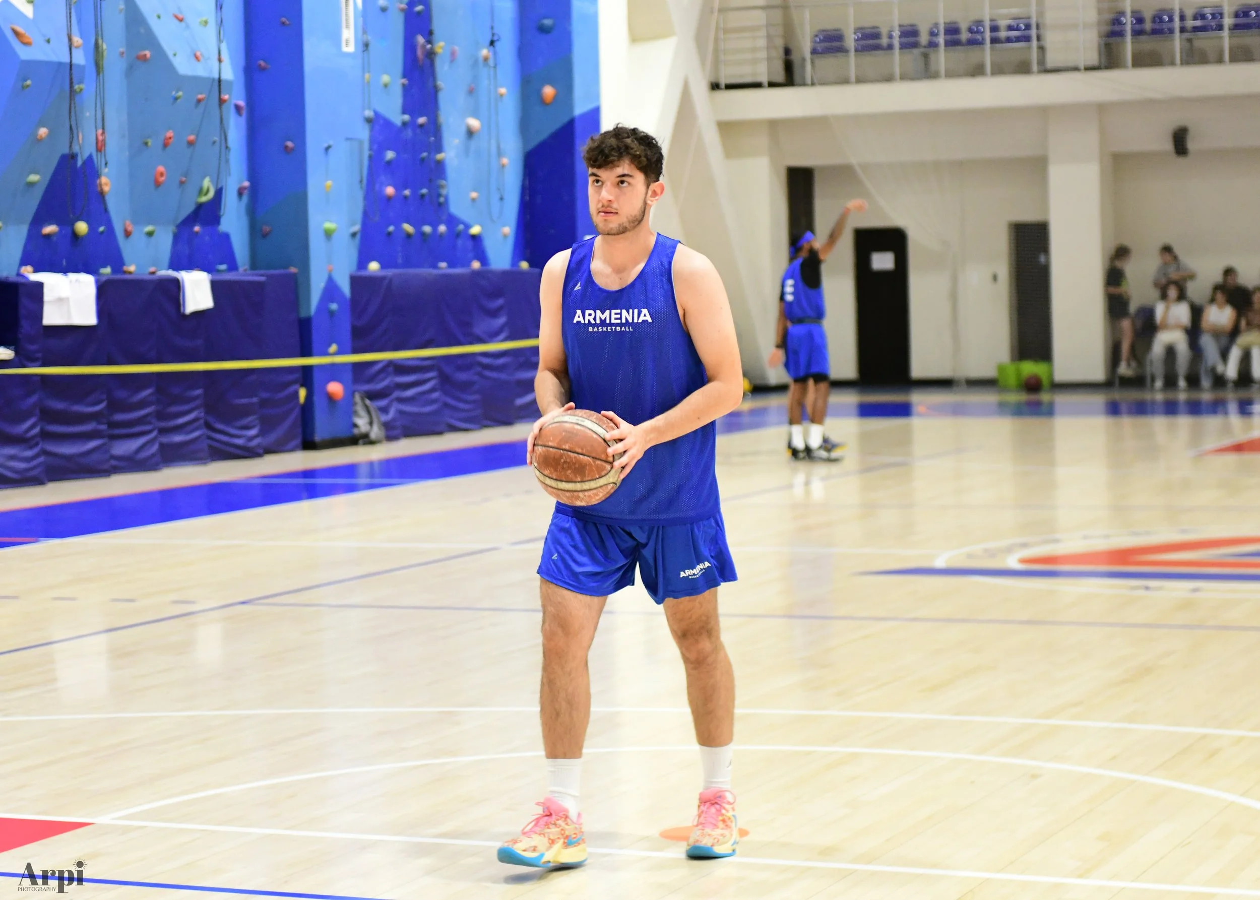 A person in a blue "Armenia Basketball" uniform holding a basketball on an indoor court. Another player in the background also in blue. The court has a climbing wall on the side.