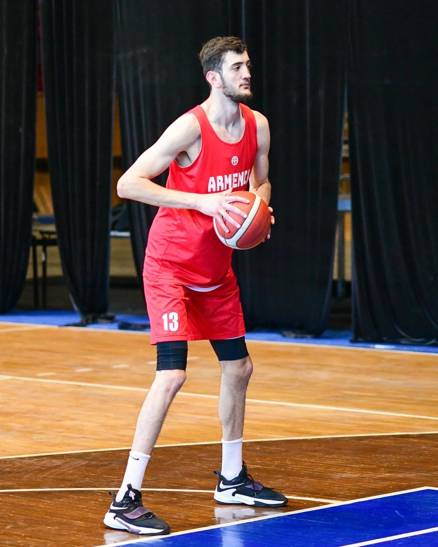 A male basketball player in a red jersey with "Armenia" on it holds a basketball on a court.