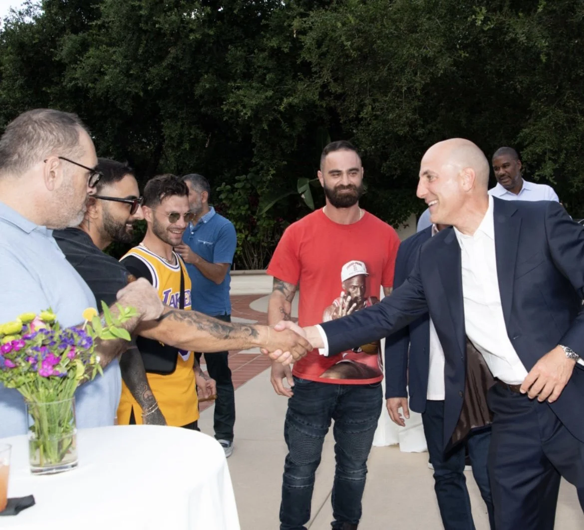 Group of people outdoors, some shaking hands and others observing, with a table and floral arrangement in the foreground.