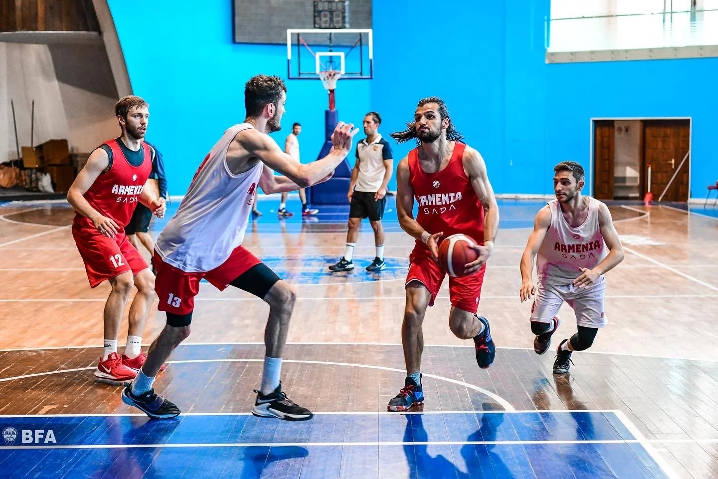 Basketball players on a court during a game or practice. Players wearing red and white jerseys with 'Armenia' printed on them. One player dribbling the ball, with others engaged in action around him. Indoor basketball court with wooden flooring and a
