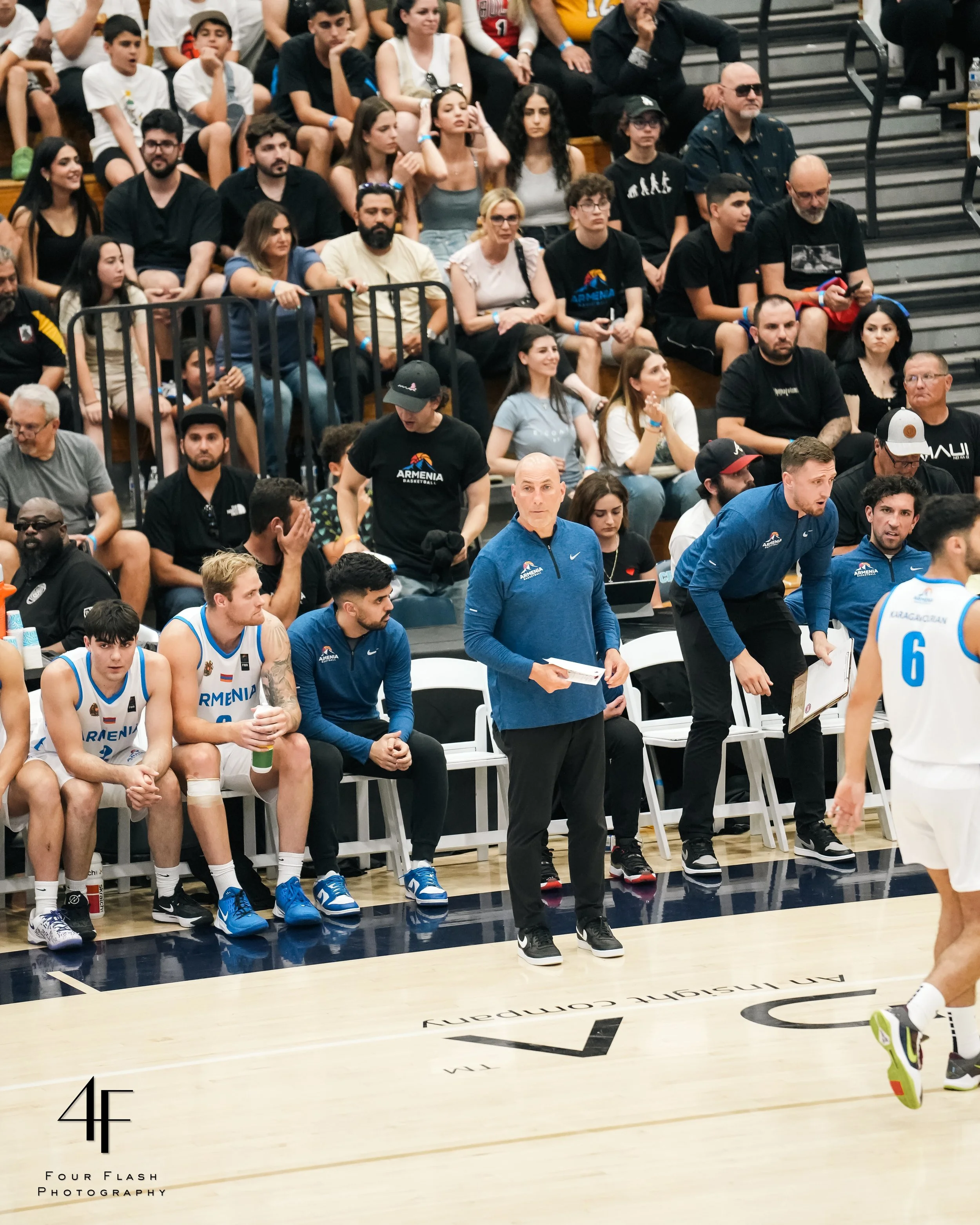 Basketball game scene with a team in white jerseys sitting on the bench, alongside coaches standing near the sideline. Spectators are visible in the bleachers.