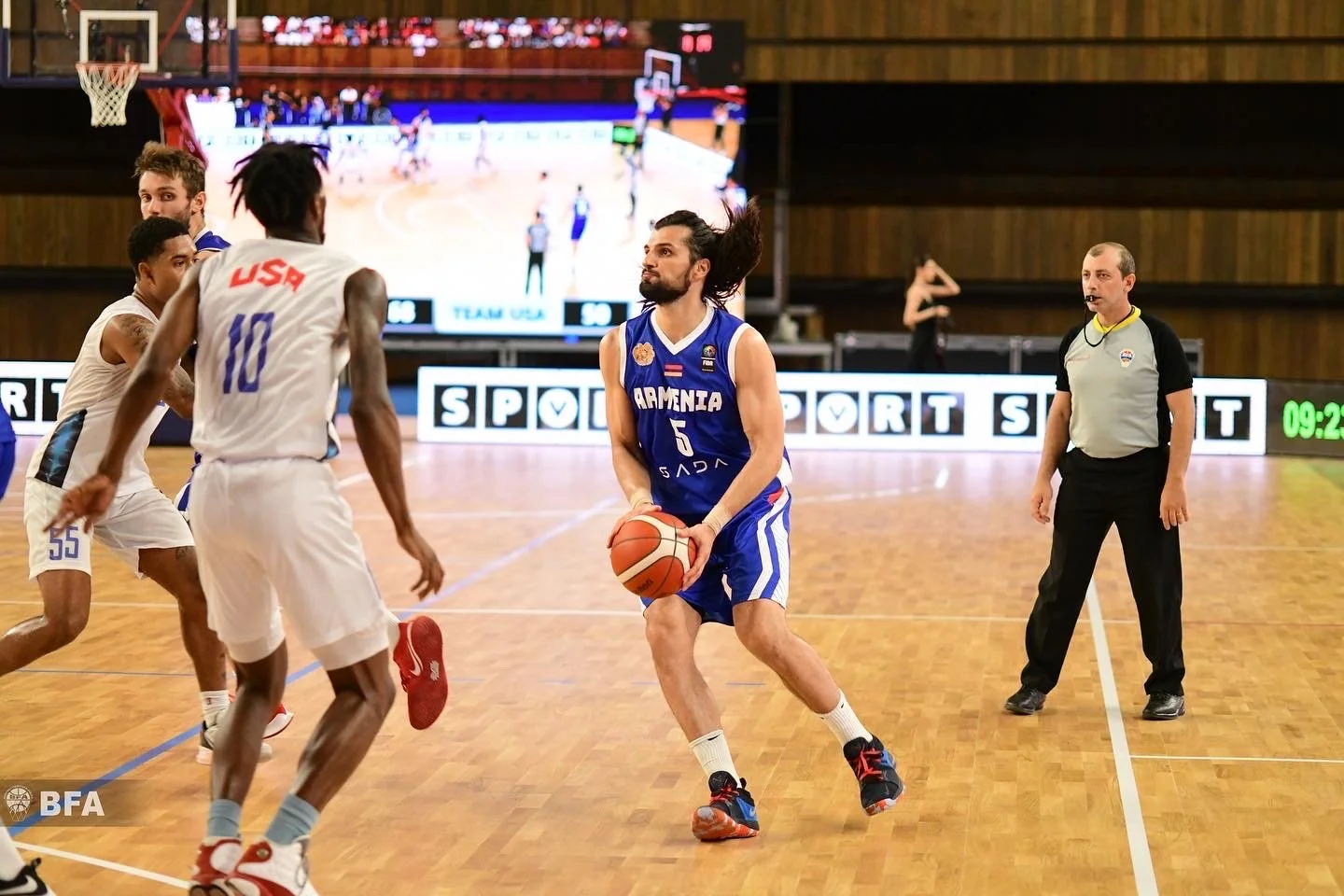 Basketball game with players from Armenia and USA; Armenian player holding the ball, USA players in white uniforms; referee observing.