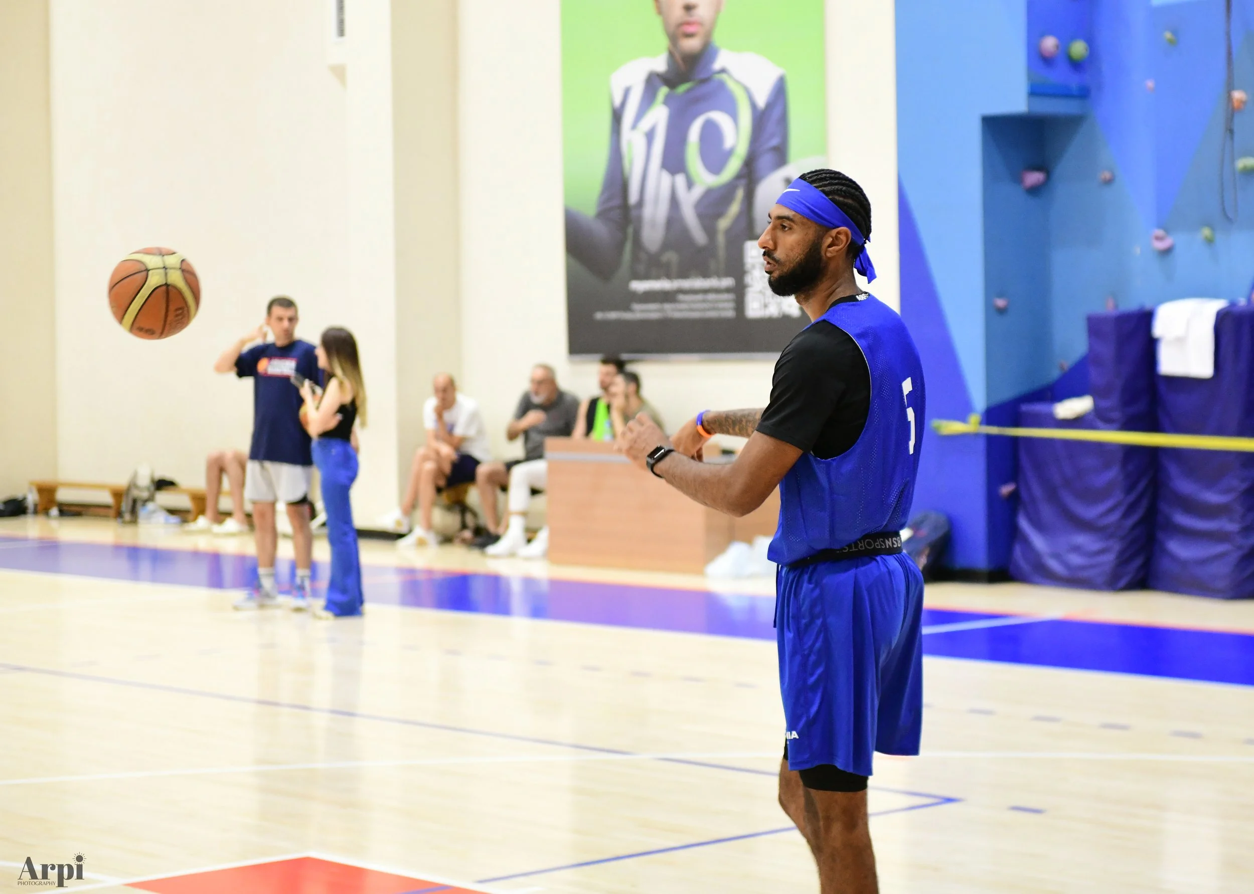 A basketball player in a blue jersey labeled with the number 5, holding a basketball on a court with people in the background. A large poster is visible on the wall.