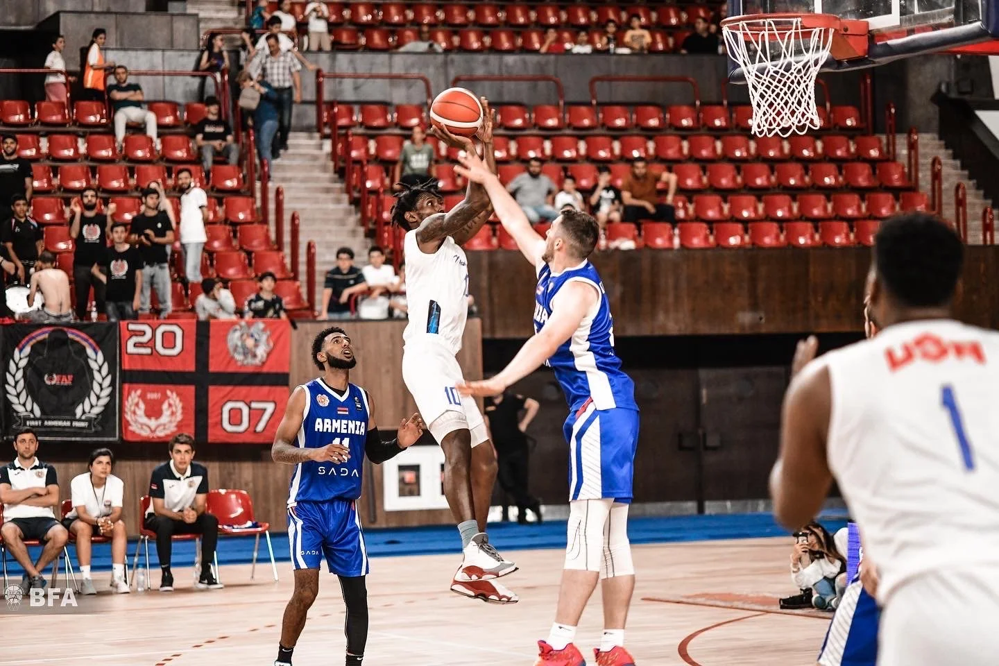 Basketball game with players in blue and white uniforms on an indoor court, one player taking a jump shot.
