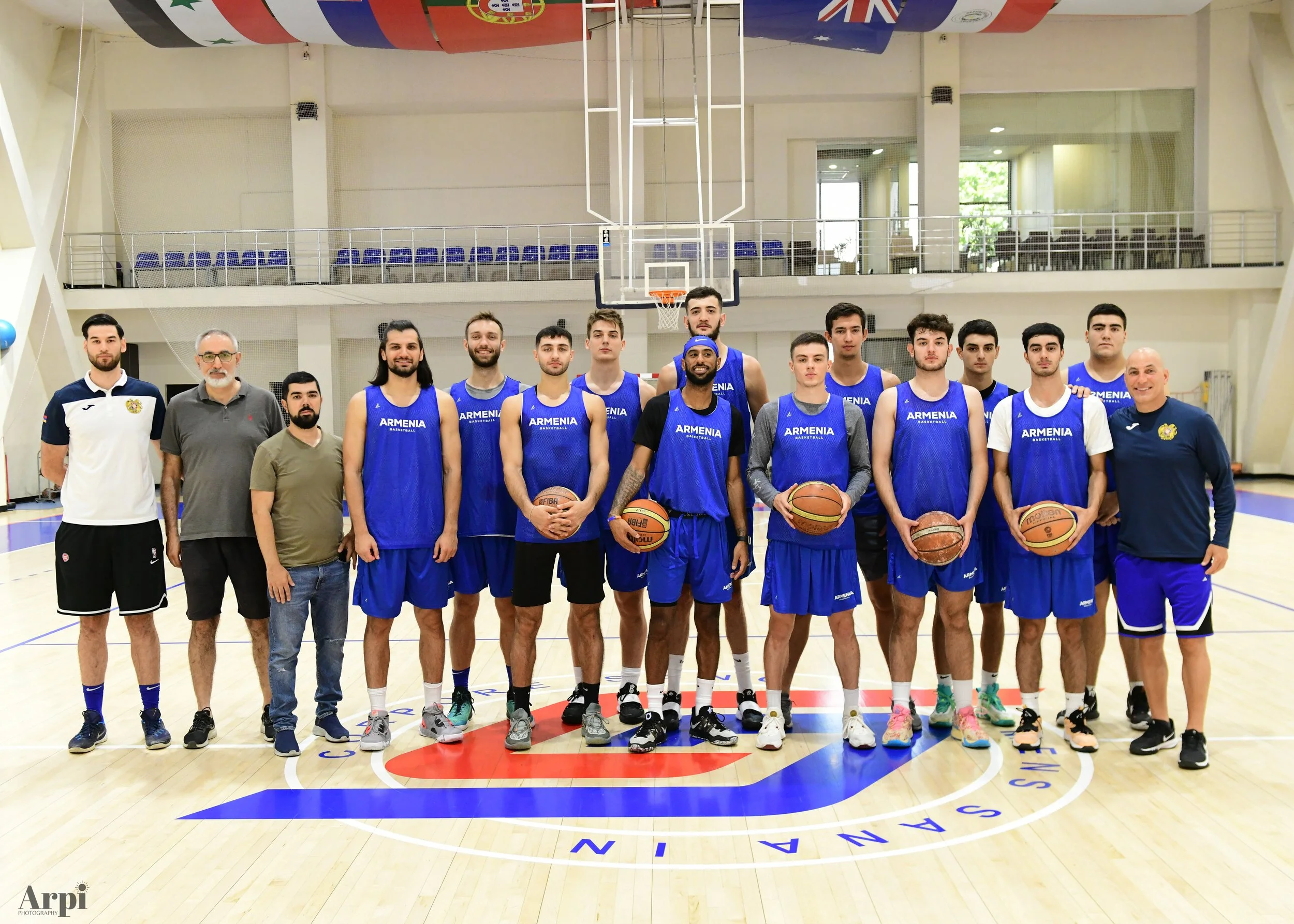 Group photo of male basketball team wearing blue "Armenia" jerseys, standing on a basketball court with flags in the background.