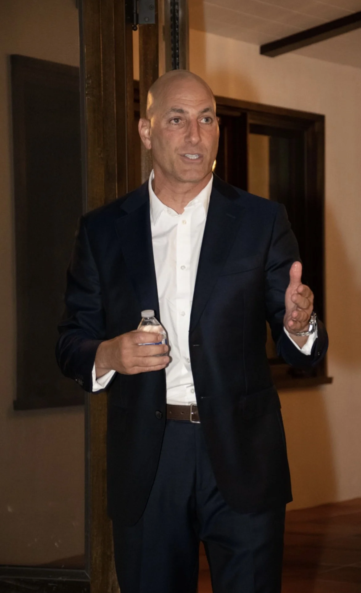 Bald man in a dark suit and white shirt, holding a water bottle, gesturing with one hand, indoors.