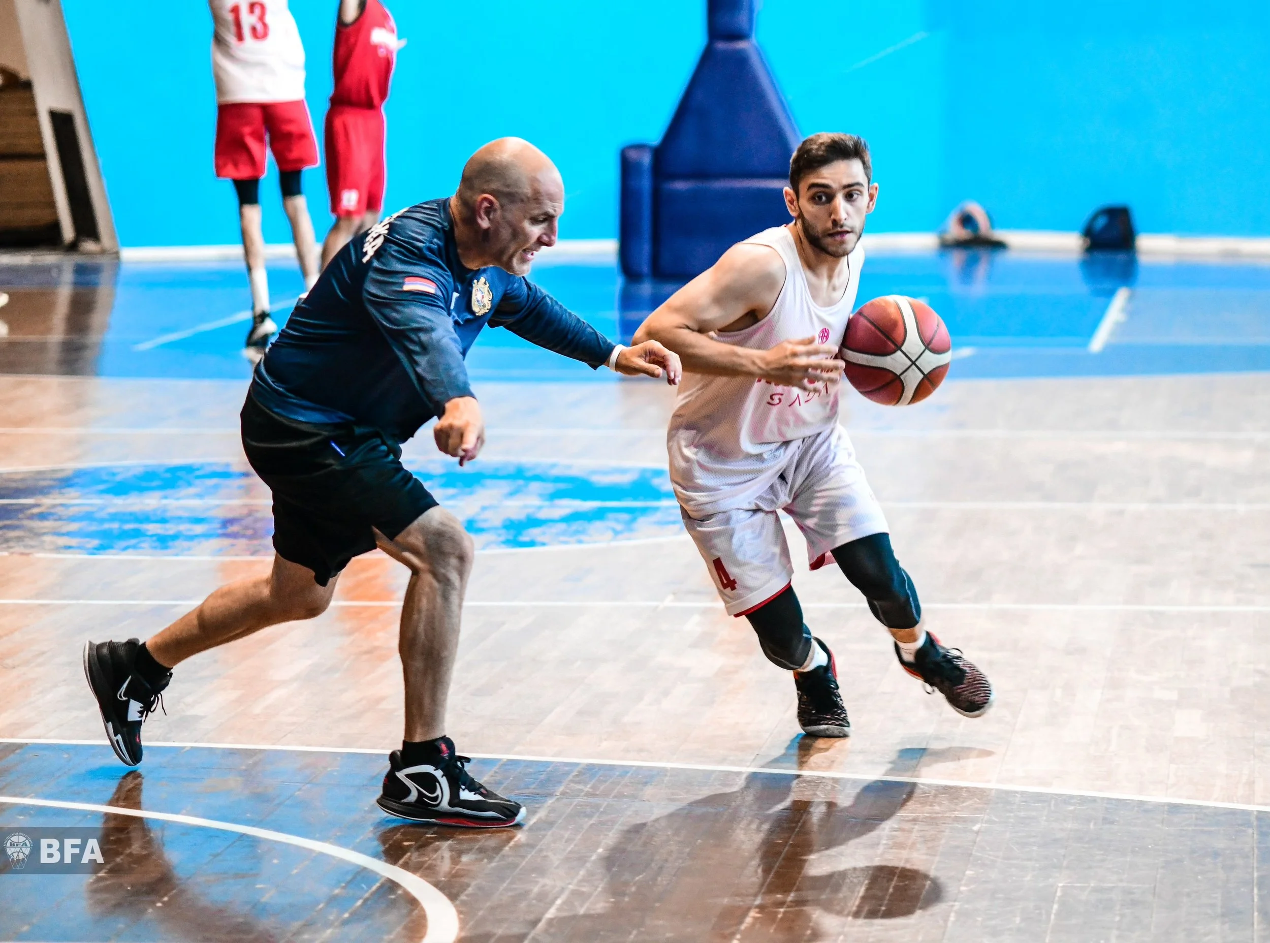 A basketball player in a white uniform dribbling past a defender in a blue jersey during a game, with other players visible in the background on an indoor court.