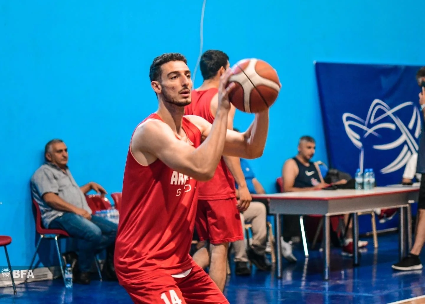 Basketball player in red jersey, number 14, preparing to shoot a basketball indoors, with a blue background and seated spectators.