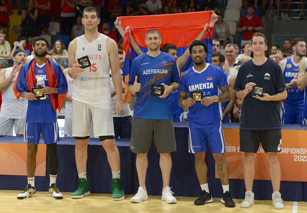 Group of basketball players holding awards, standing in front of a sports event banner, with a crowd in the background. Players have flags draped over shoulders.