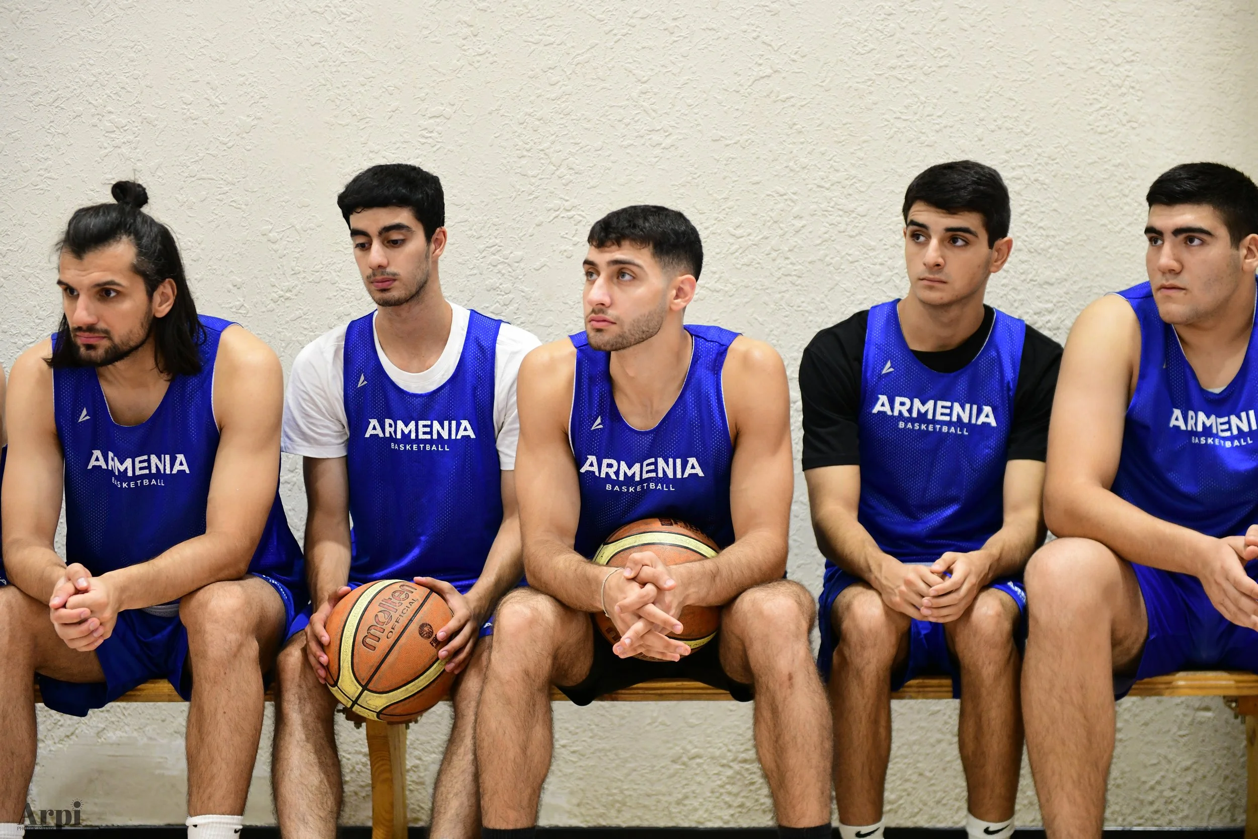 Five basketball players wearing blue sleeveless jerseys with 'Armenia Basketball' printed on them, sitting on a bench, holding basketballs.