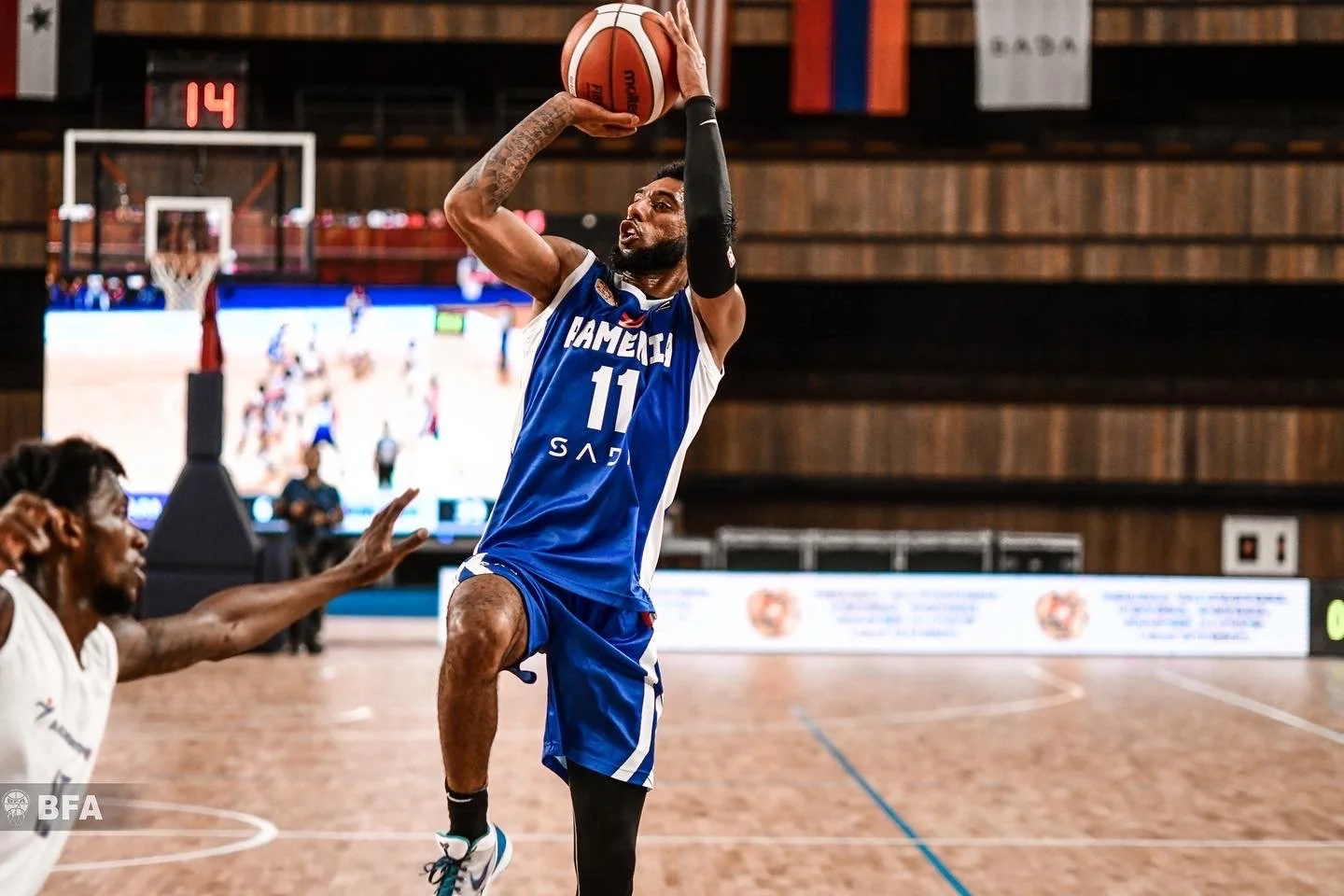 Basketball player in blue jersey jumps for a shot during a game, with another player in white reaching to block. Court and scoreboard visible in the background.