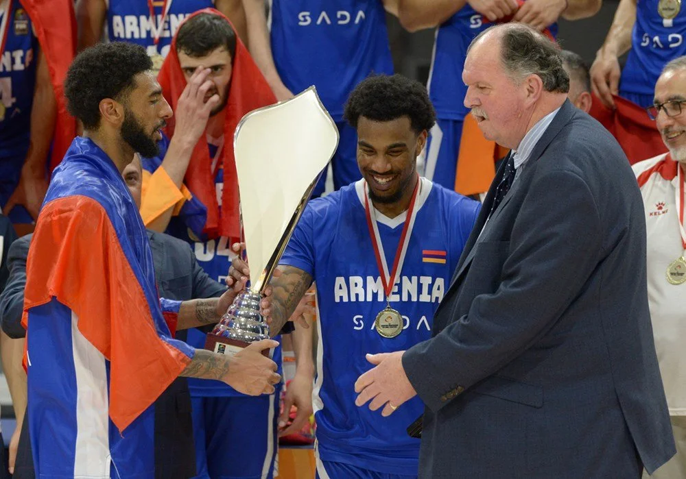 Basketball players in Armenia jerseys holding a trophy, celebrating victory. One player is draped in an Armenian flag, and a medal is visible on another player's neck. A man in a suit is presenting the trophy.