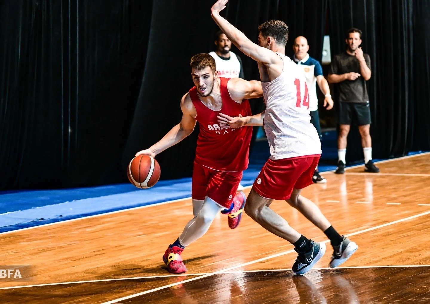Two male basketball players in a game, one wearing a red jersey with "SA" visible, dribbling the ball past a defender in a white jersey with the number 14 on it. Both are on a wooden court with a black curtain backdrop and several people observing in