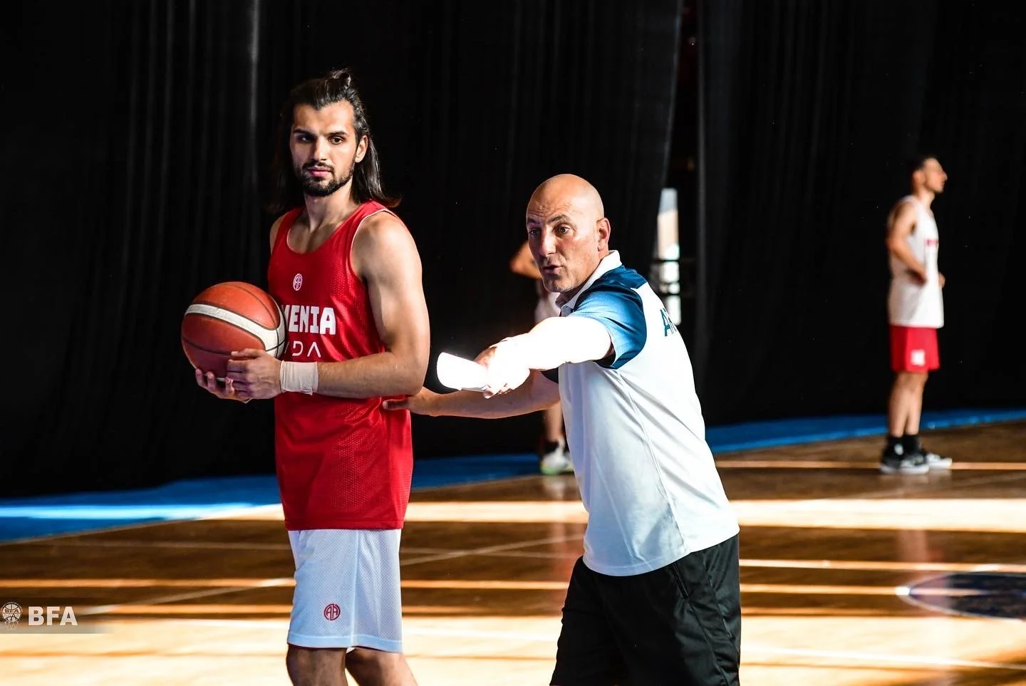Basketball player in red Slovenia jersey holding a basketball, with a coach instructing nearby on a court.