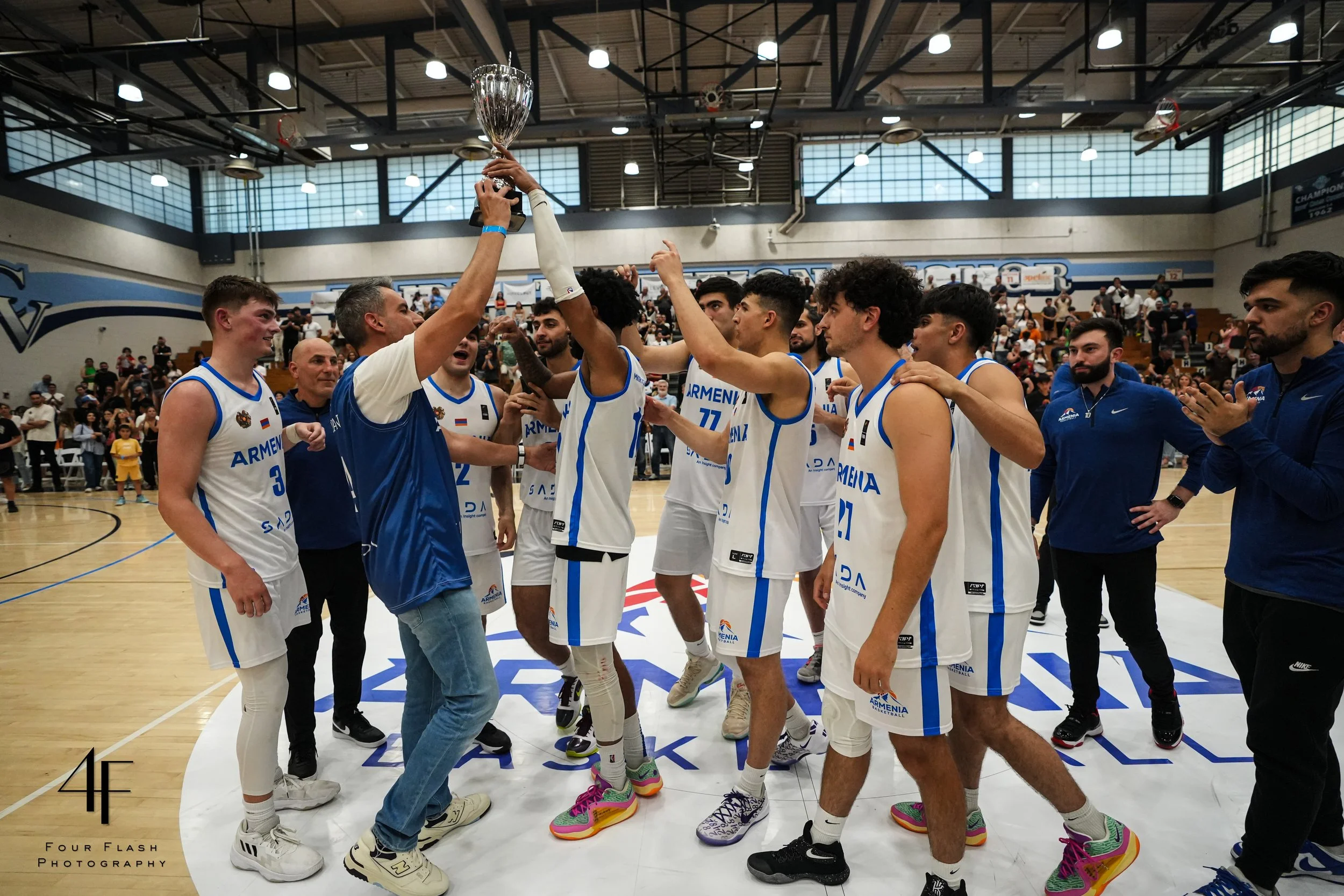 Basketball team celebrating with a trophy on the court, wearing white uniforms with 'Armenia' text, surrounded by onlookers in a gymnasium. Photography credit to Four Flash Photography.