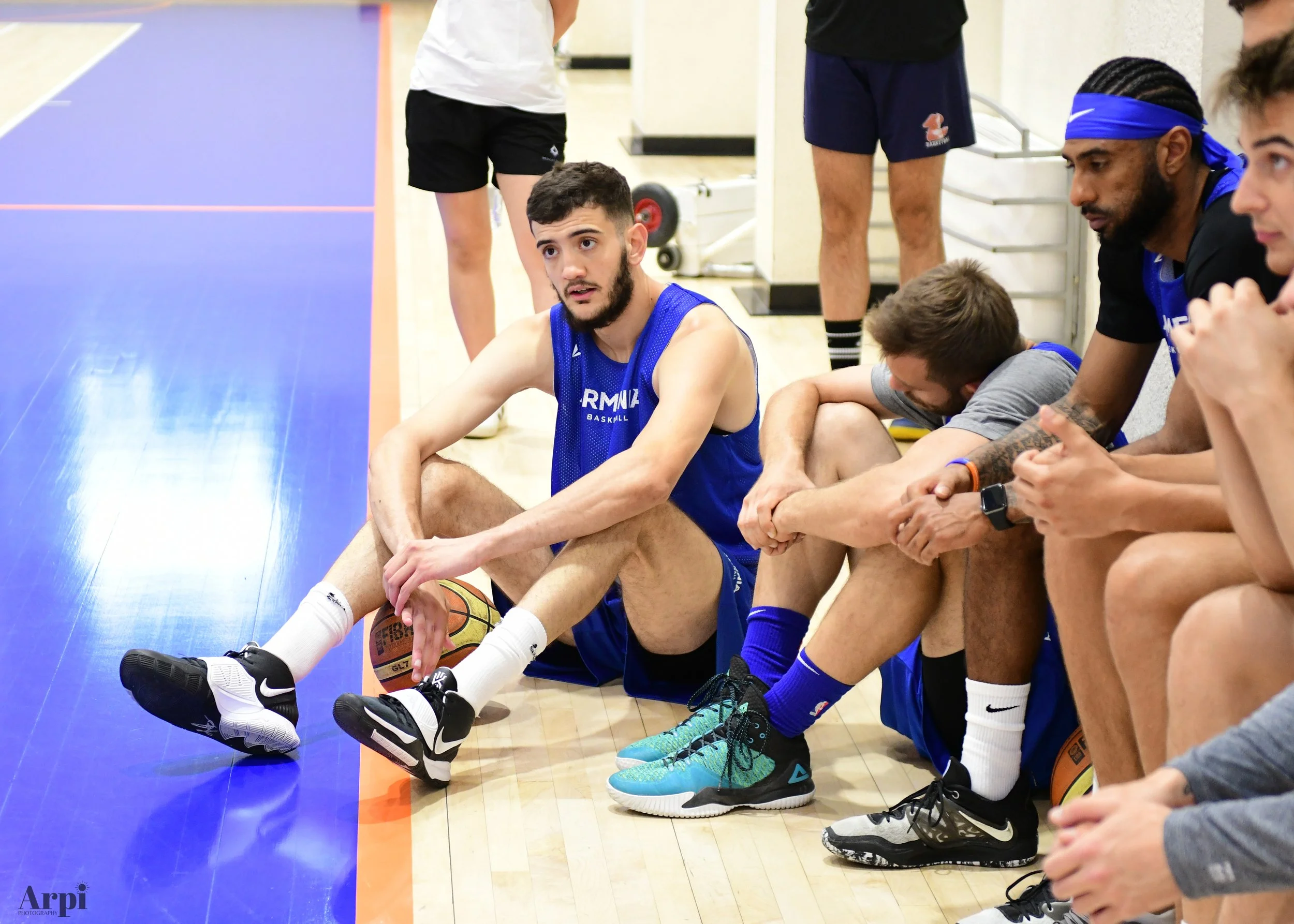 Basketball players sitting courtside in a gym during practice, with one holding a basketball. They appear to be taking a break, wearing sports attire including jerseys and sneakers.