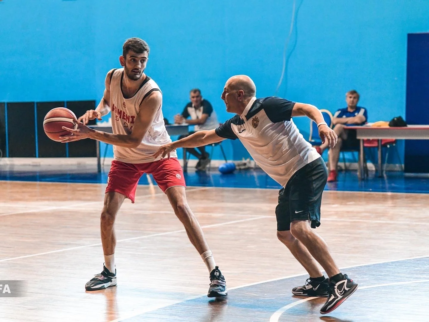 Two basketball players practicing on an indoor court, one dribbling the ball while the other defends, with observers in the background.