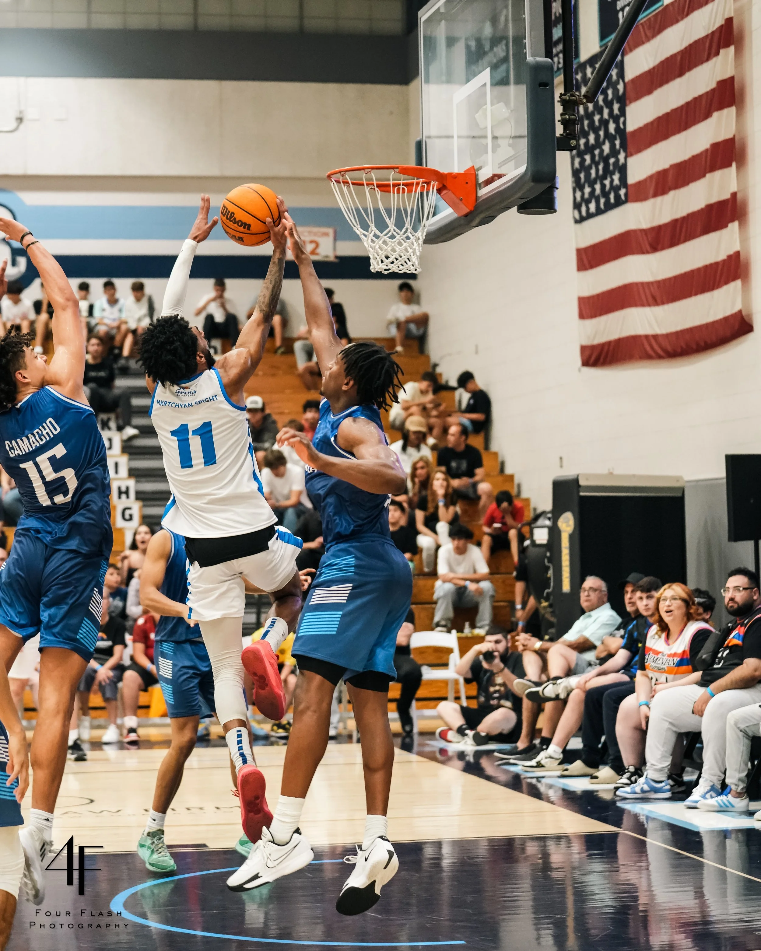 Basketball players in action beneath a hoop during a game, with an audience in the background and an American flag hanging on the wall.