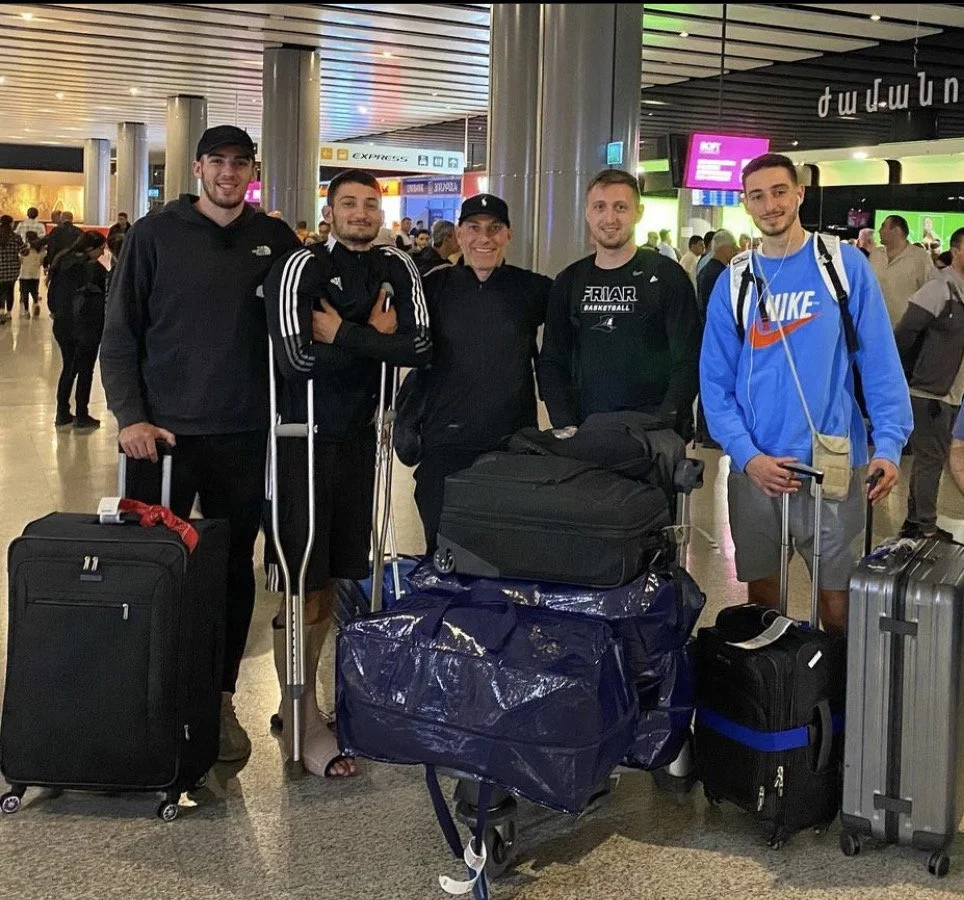 Group of five men at an airport with luggage, one on crutches.