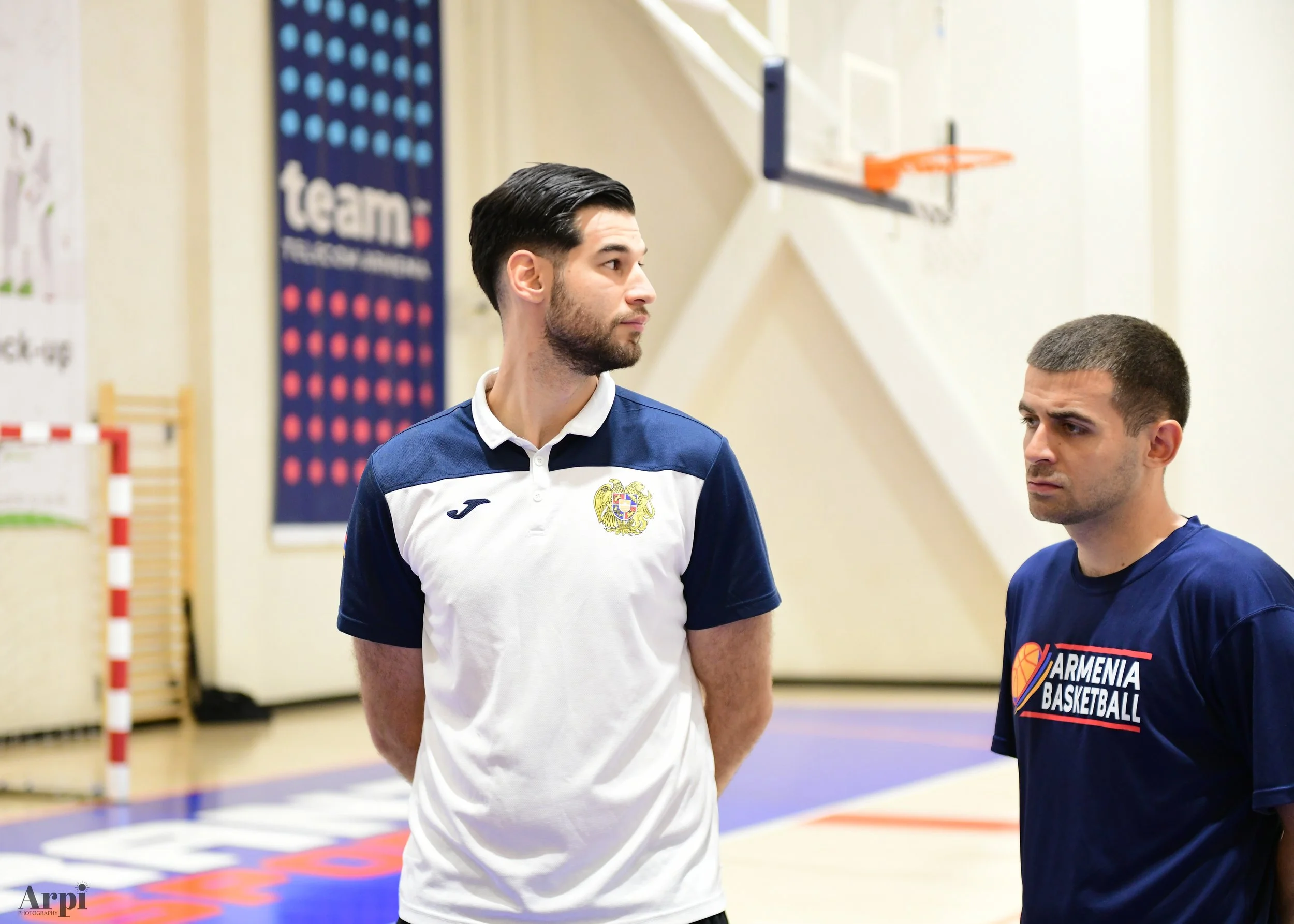 Two men standing in a gymnasium; one wearing a jersey with an Armenia crest and the other with 'Armenia Basketball' on his shirt, with a basketball court and hoop in the background.