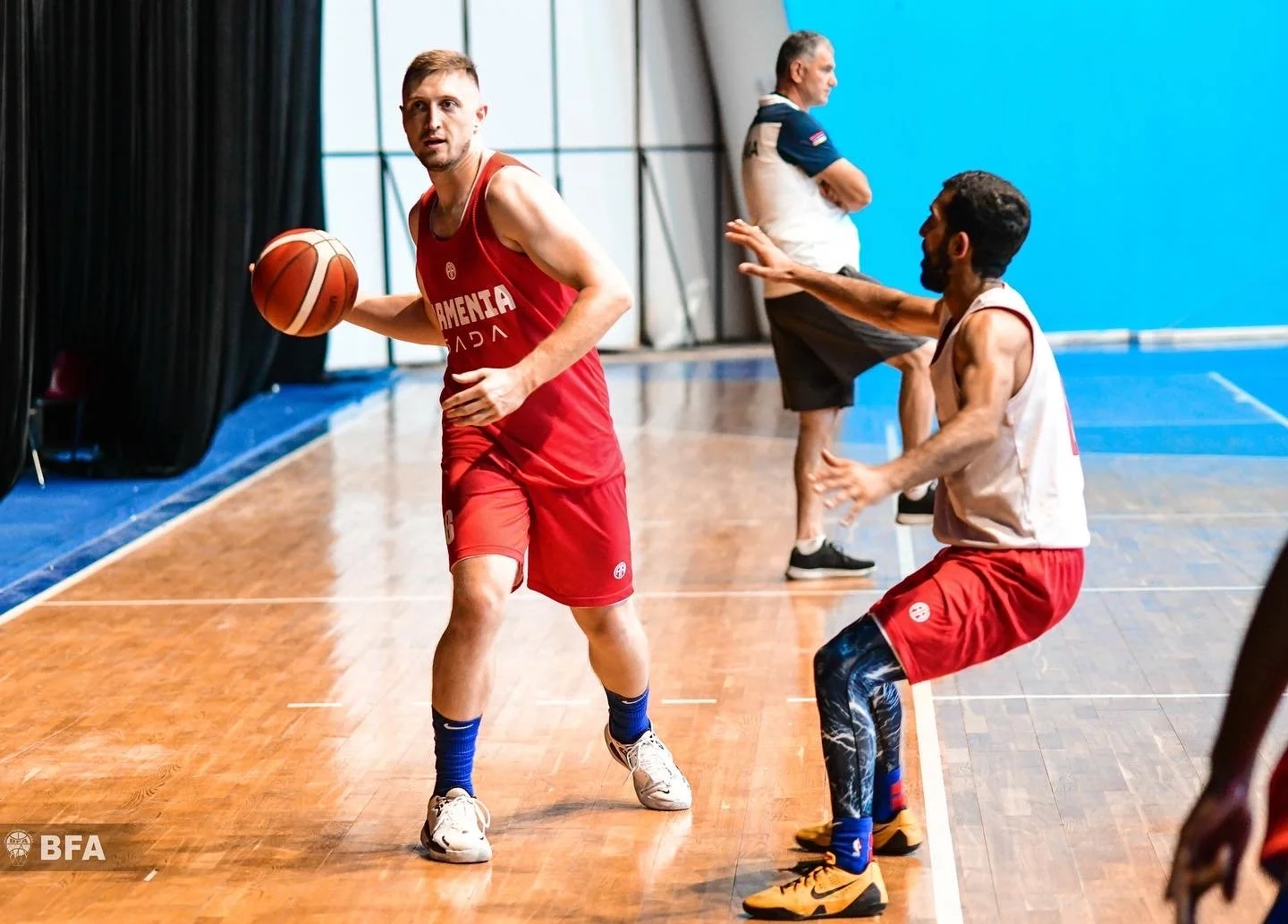 Basketball players on an indoor court, one dribbling and another defending, with a coach observing the play. Players wearing red and white uniforms.