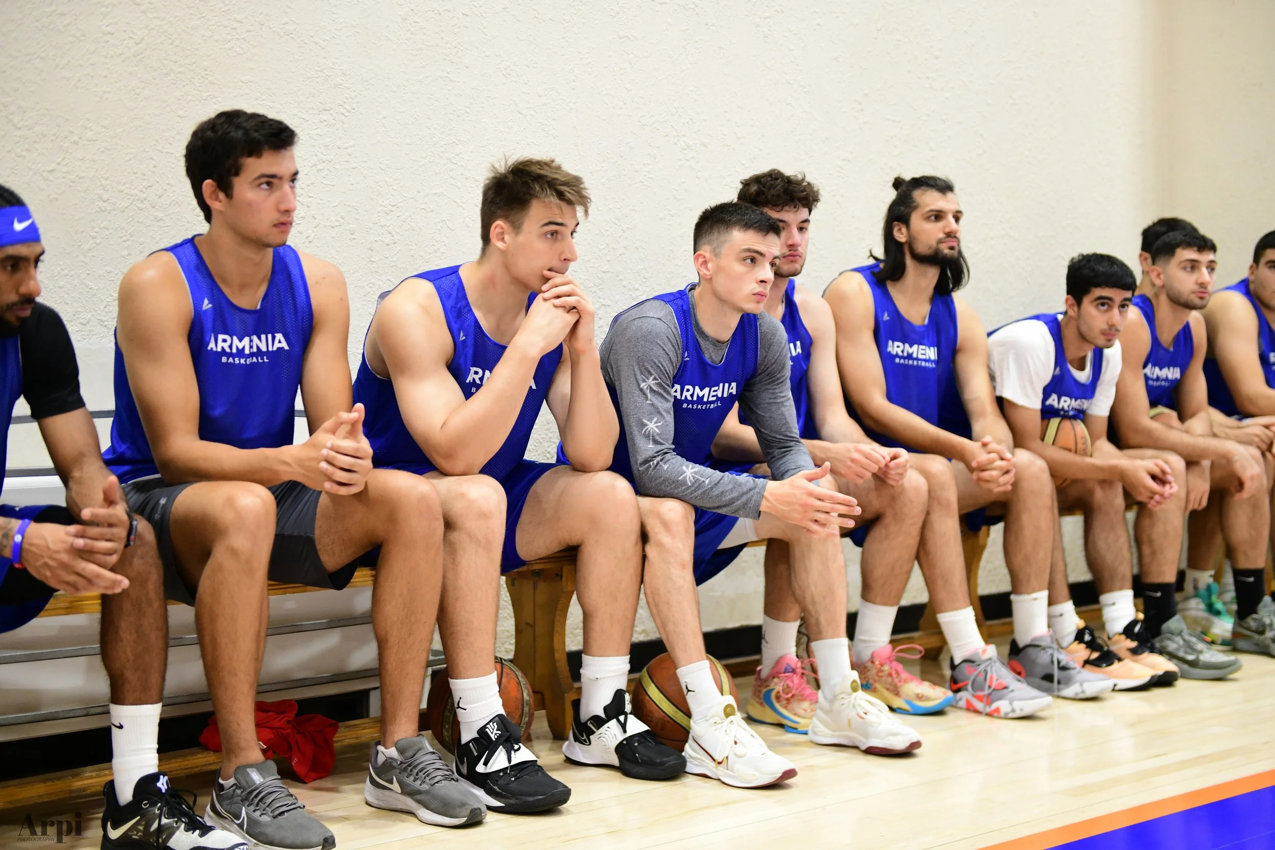 Basketball team players from Armenia sitting on a bench in blue jerseys during practice.