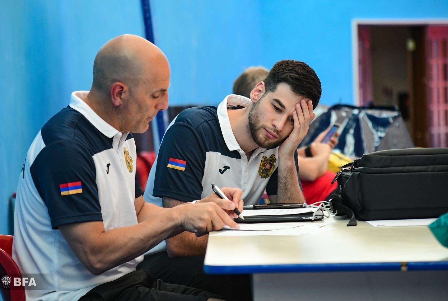 Two men in sports uniforms sitting at a desk, reviewing documents and a laptop, appearing focused and contemplative.