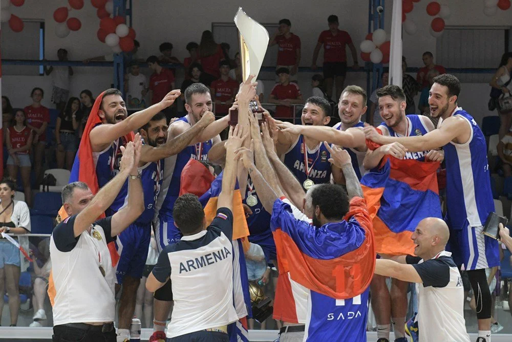 Basketball team celebrating with trophy, wearing blue and orange uniforms, holding an Armenian flag, surrounded by audience in gymnasium.