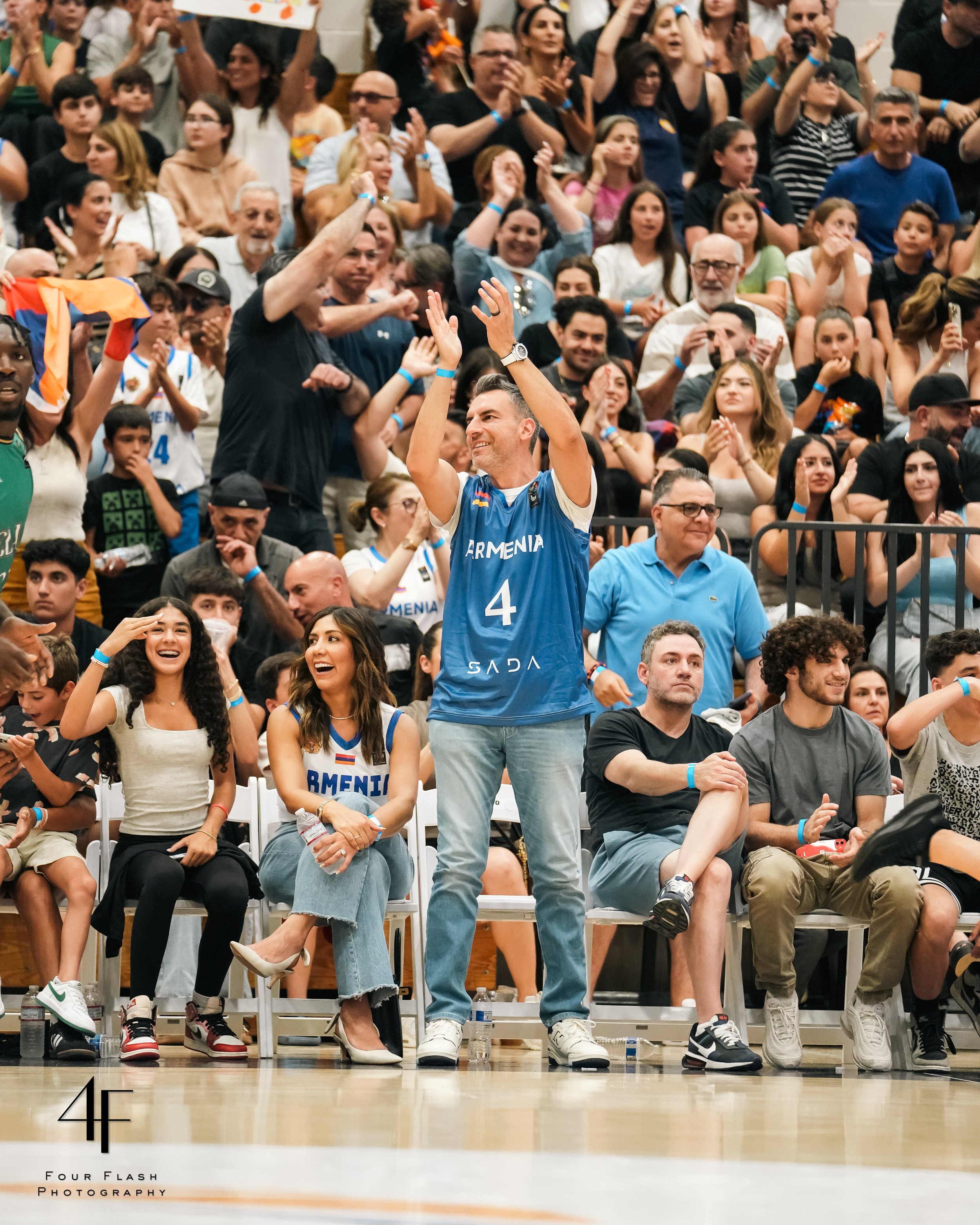 Basketball game crowd cheering, people wearing blue and white jerseys, man standing clapping, seated spectators smiling and applauding, lively atmosphere.
