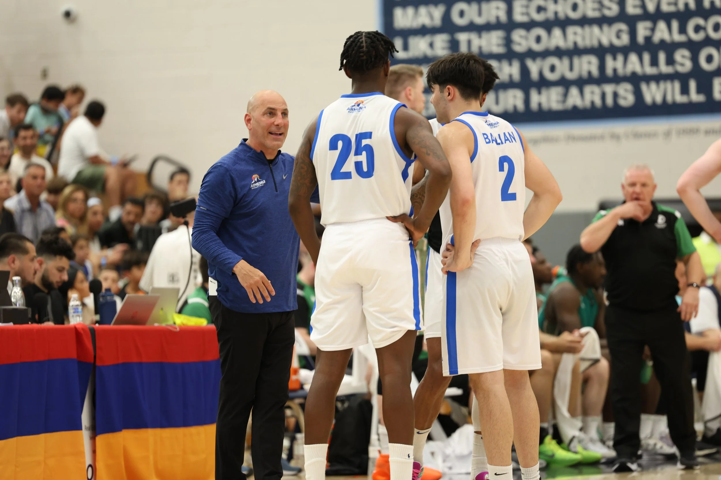 Basketball coach instructing players on sideline during a game, with audience and officials in the background.