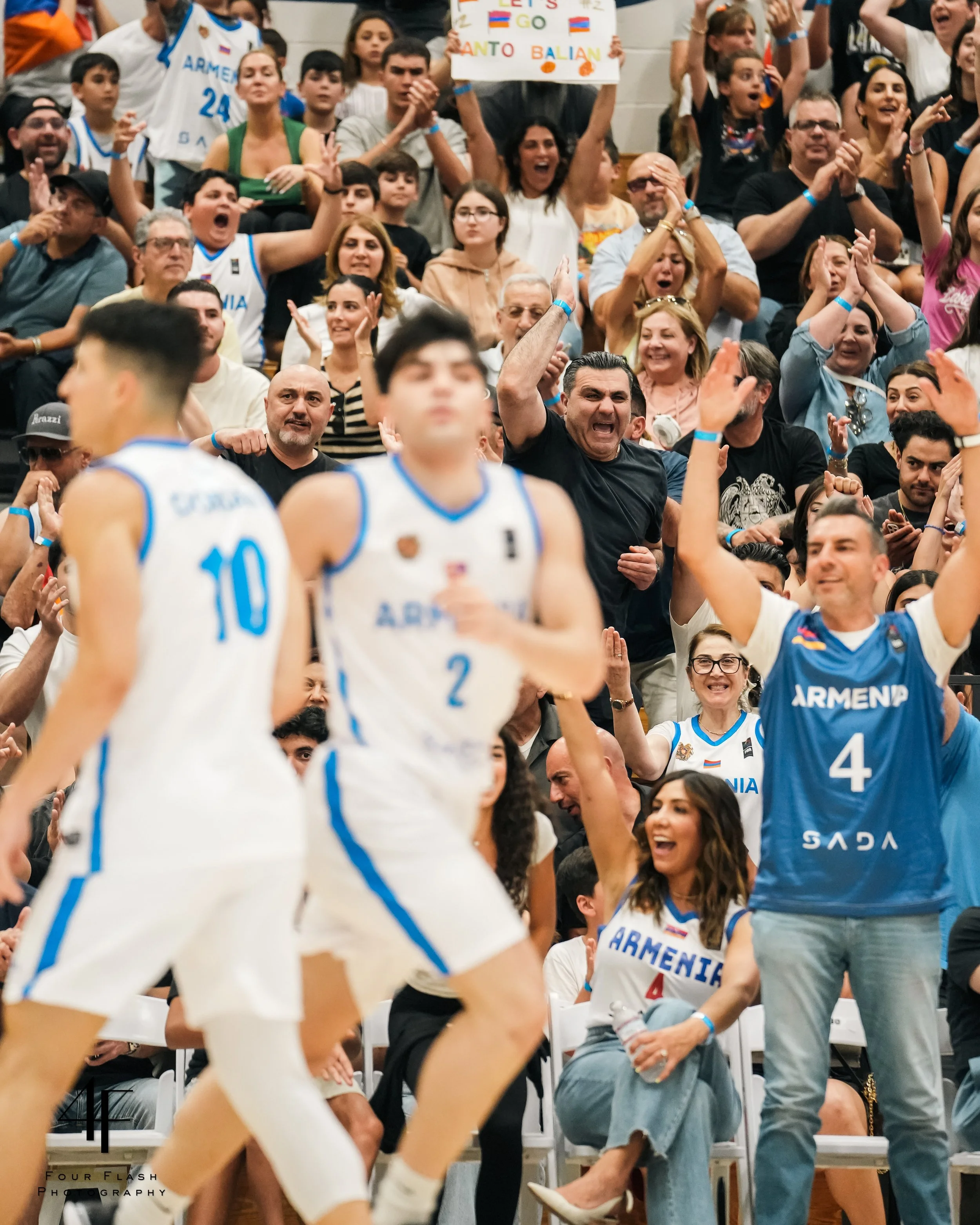 Excited fans cheering at a basketball game with players wearing Armenia jerseys. A person holds a sign saying "Let’s Go Santo Balian" in the background.