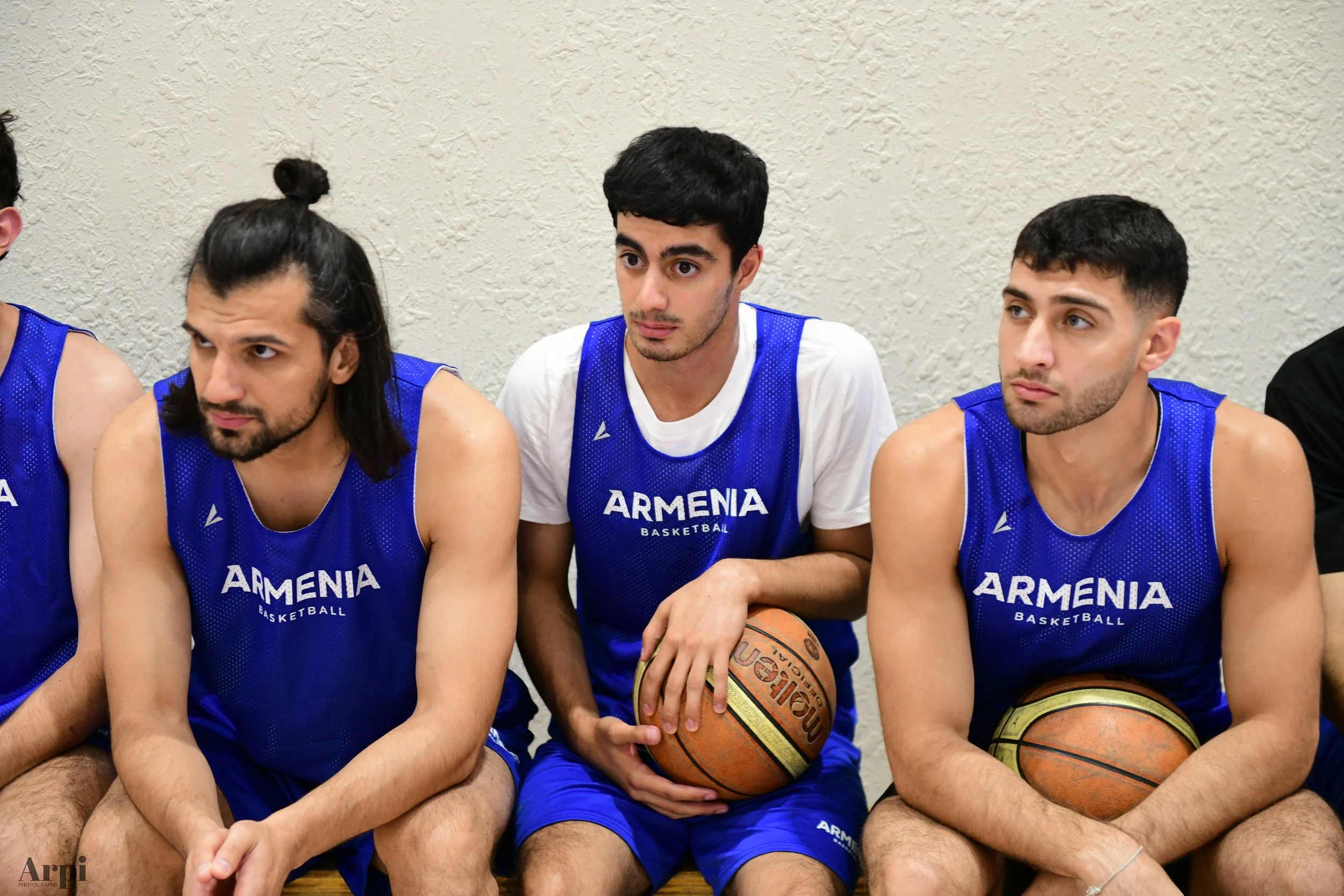 Basketball players wearing blue jerseys labeled 'Armenia' seated on a bench holding basketballs.