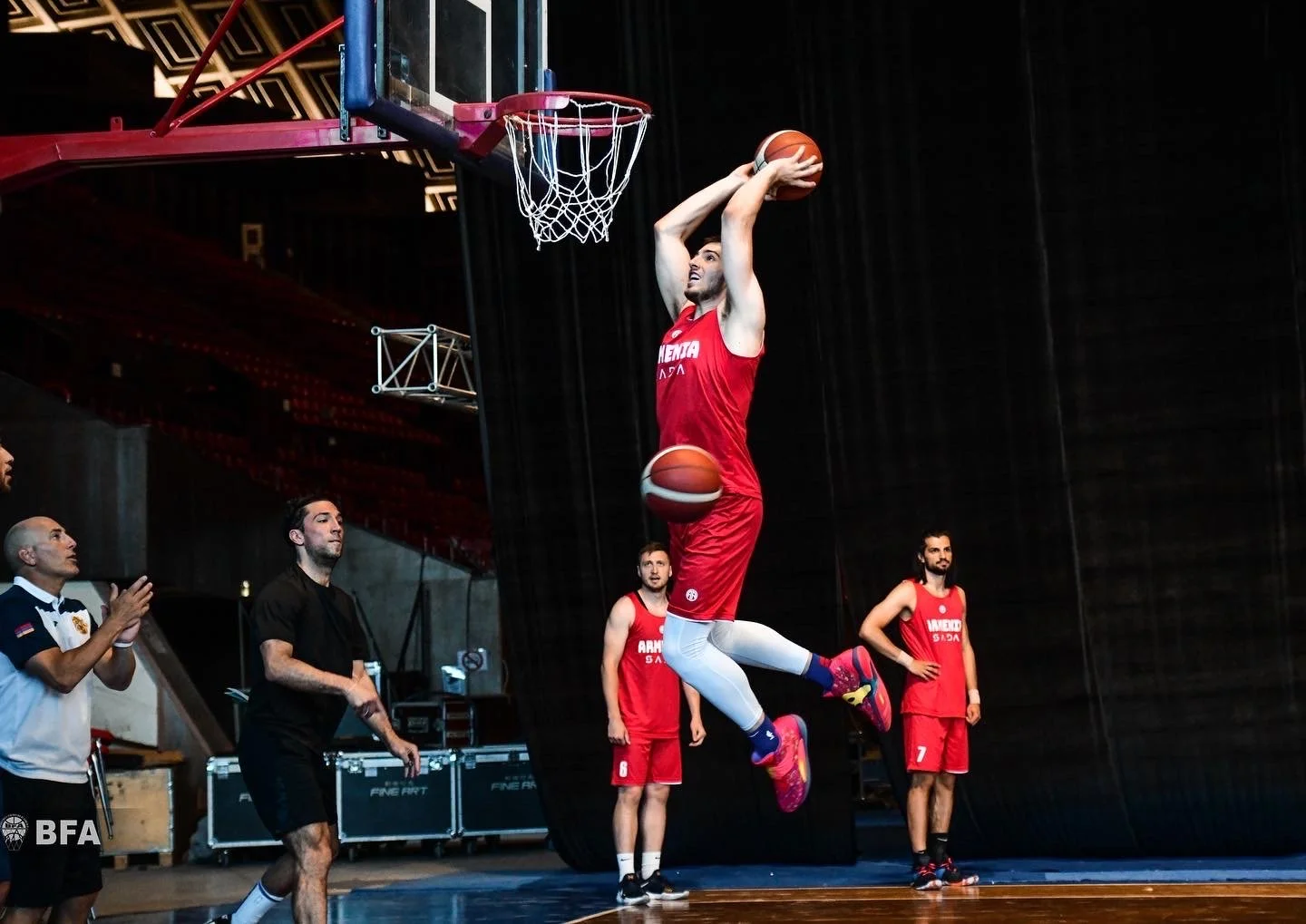 Basketball player in a red jersey jumping to dunk the ball, with spectators and teammates watching in a gym.