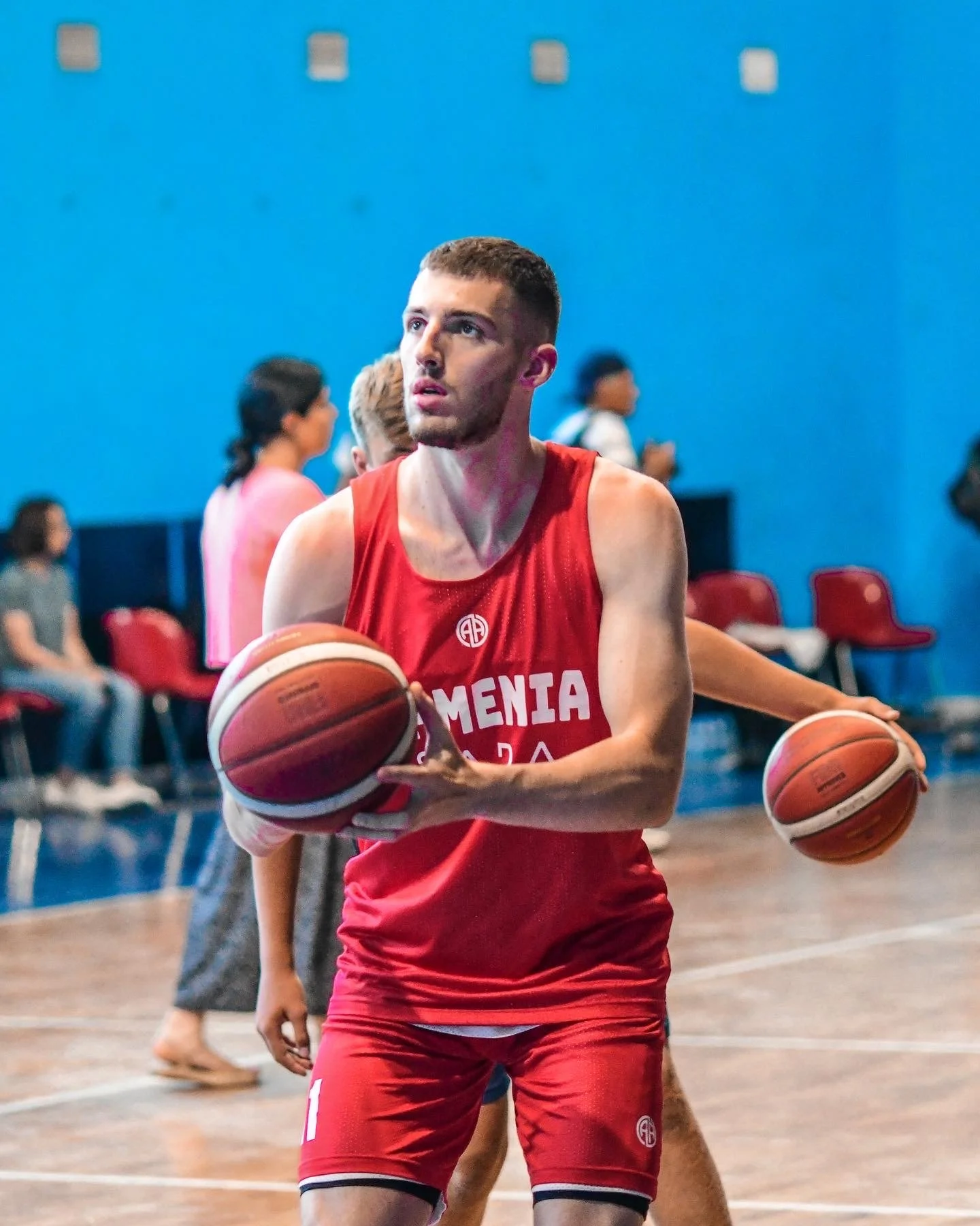 Basketball player in red uniform holding two basketballs on an indoor court