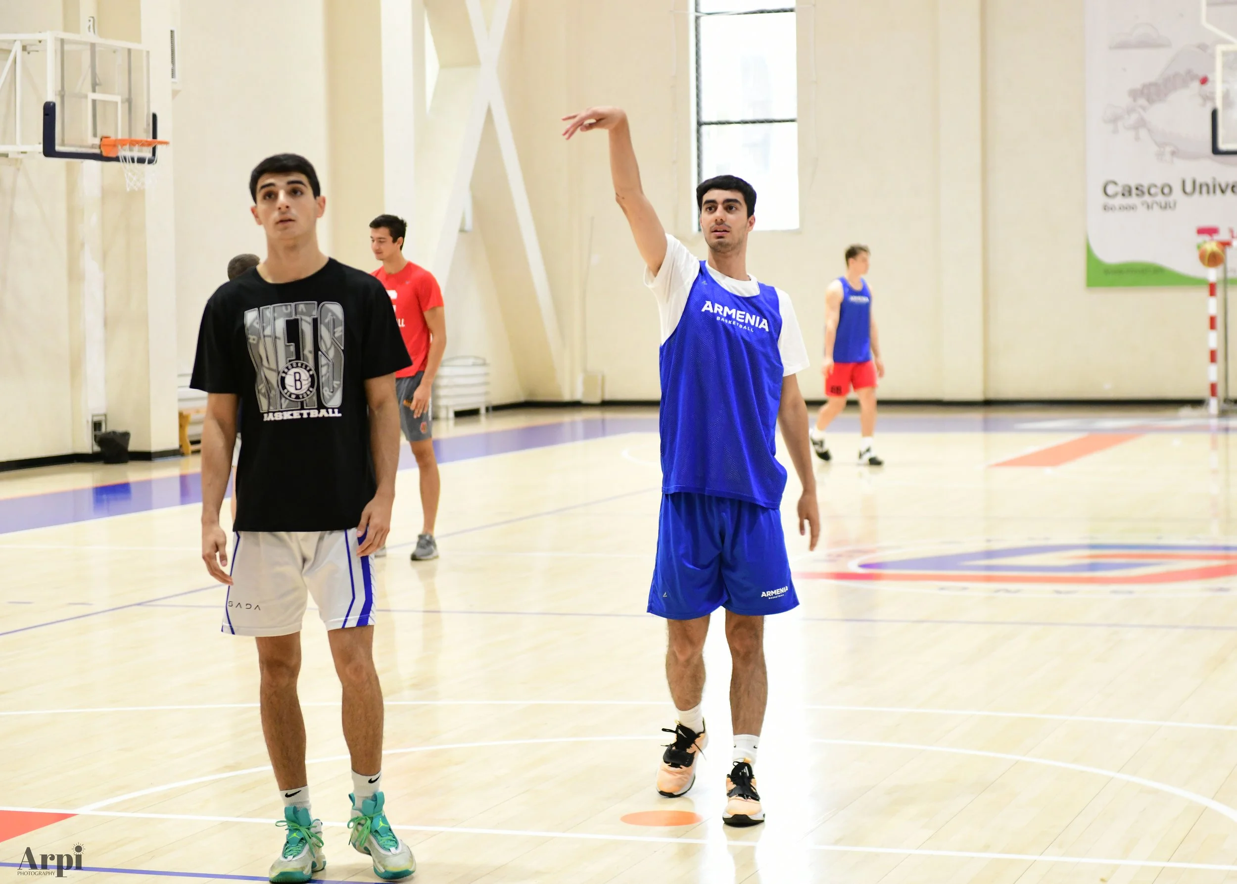 Basketball players practicing in a gym, with one player shooting and another observing.