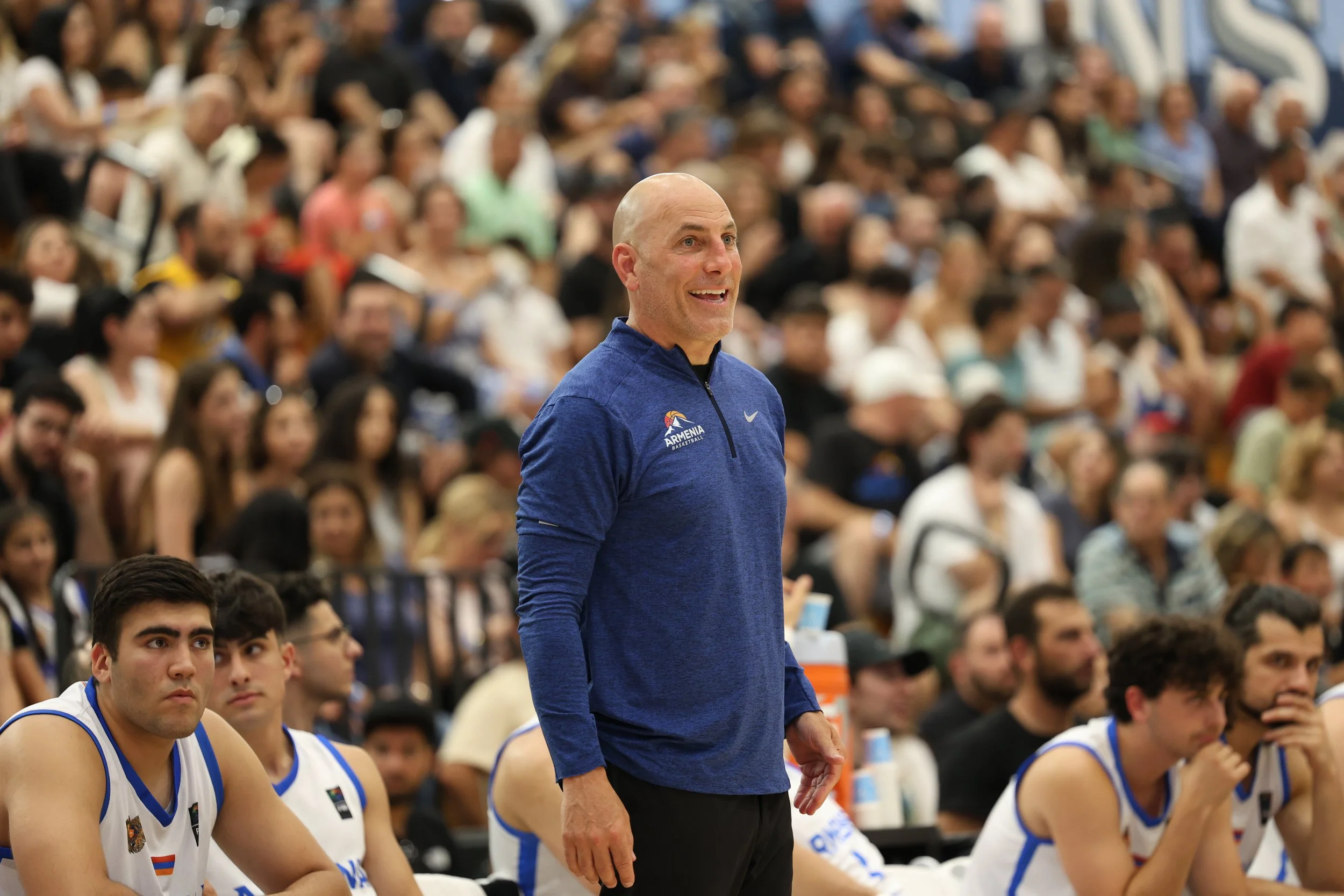 Basketball coach in blue jacket smiling, surrounded by players and spectators in a gymnasium.