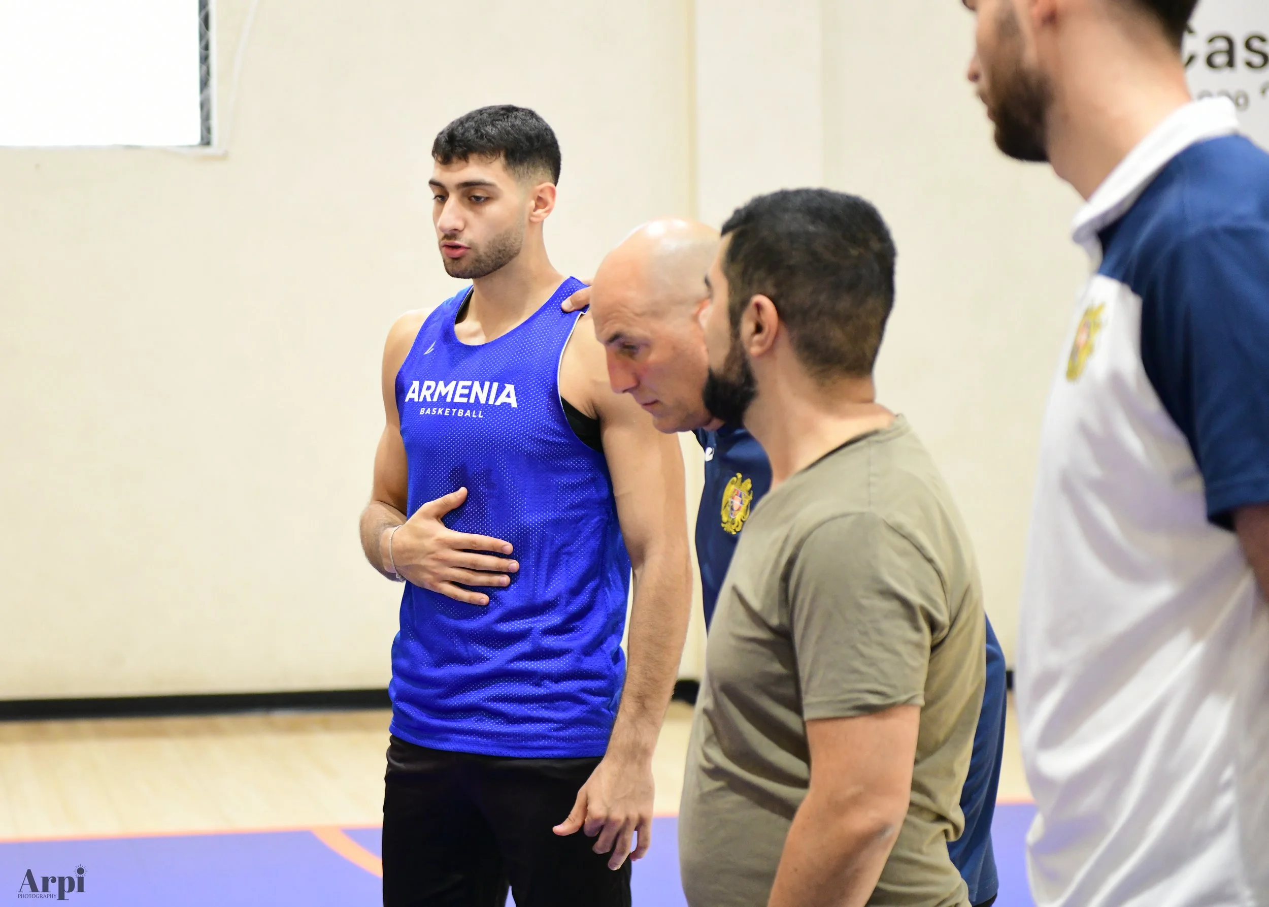 A group of men in a gym setting. One wearing a blue "Armenia Basketball" jersey, appearing to be in a discussion or a practice with others.