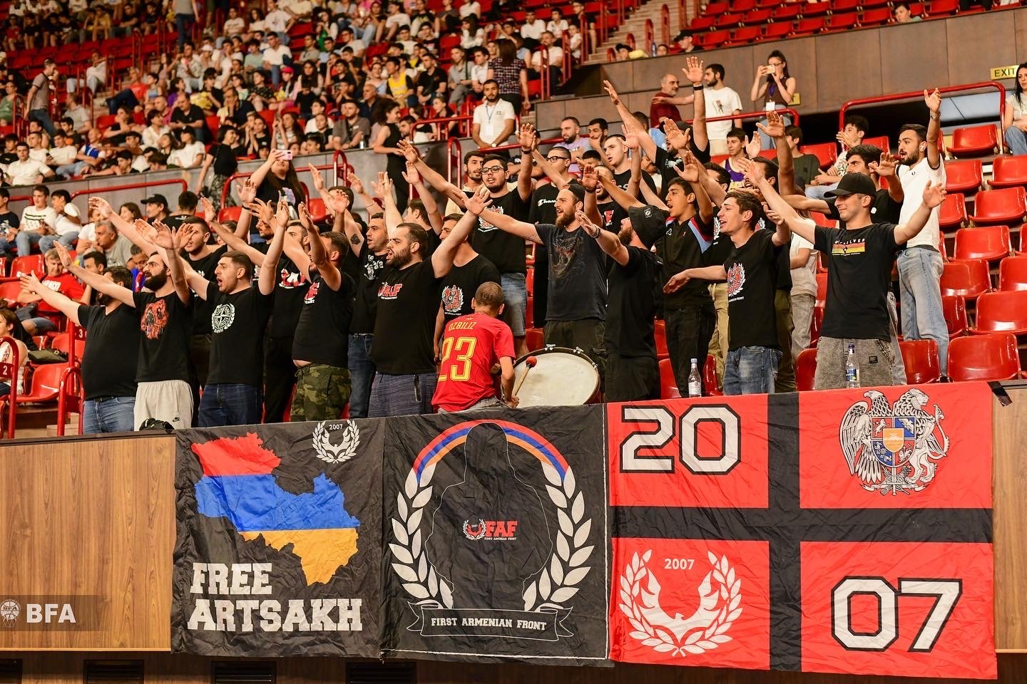 Fans cheering in a stadium with flags and banners supporting Artsakh, including "Free Artsakh" text, and First Armenian Front references.