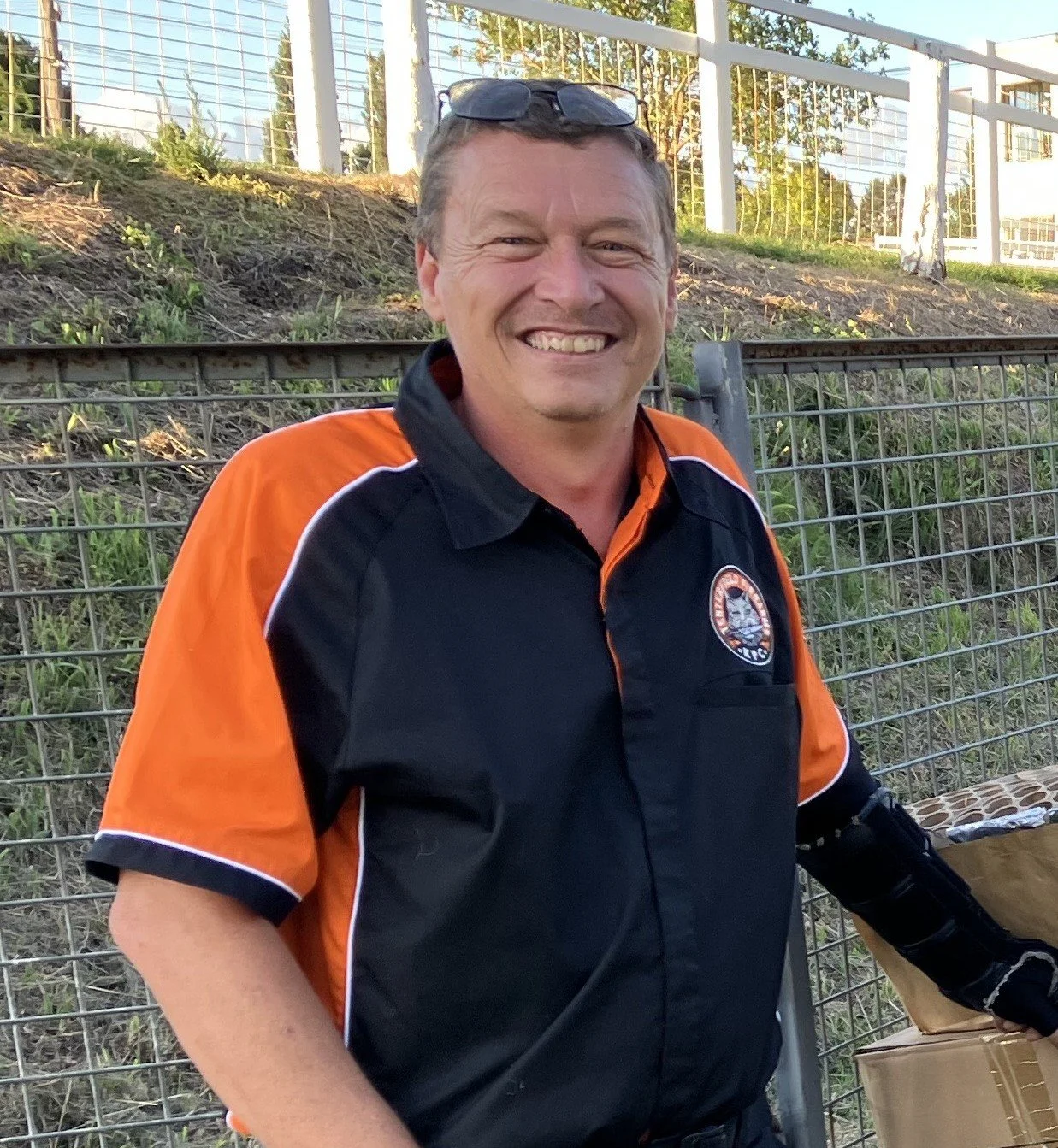 Smiling man in a black and orange shirt with a patch, standing outdoors near a wire fence, with trees and a sports field in the background.