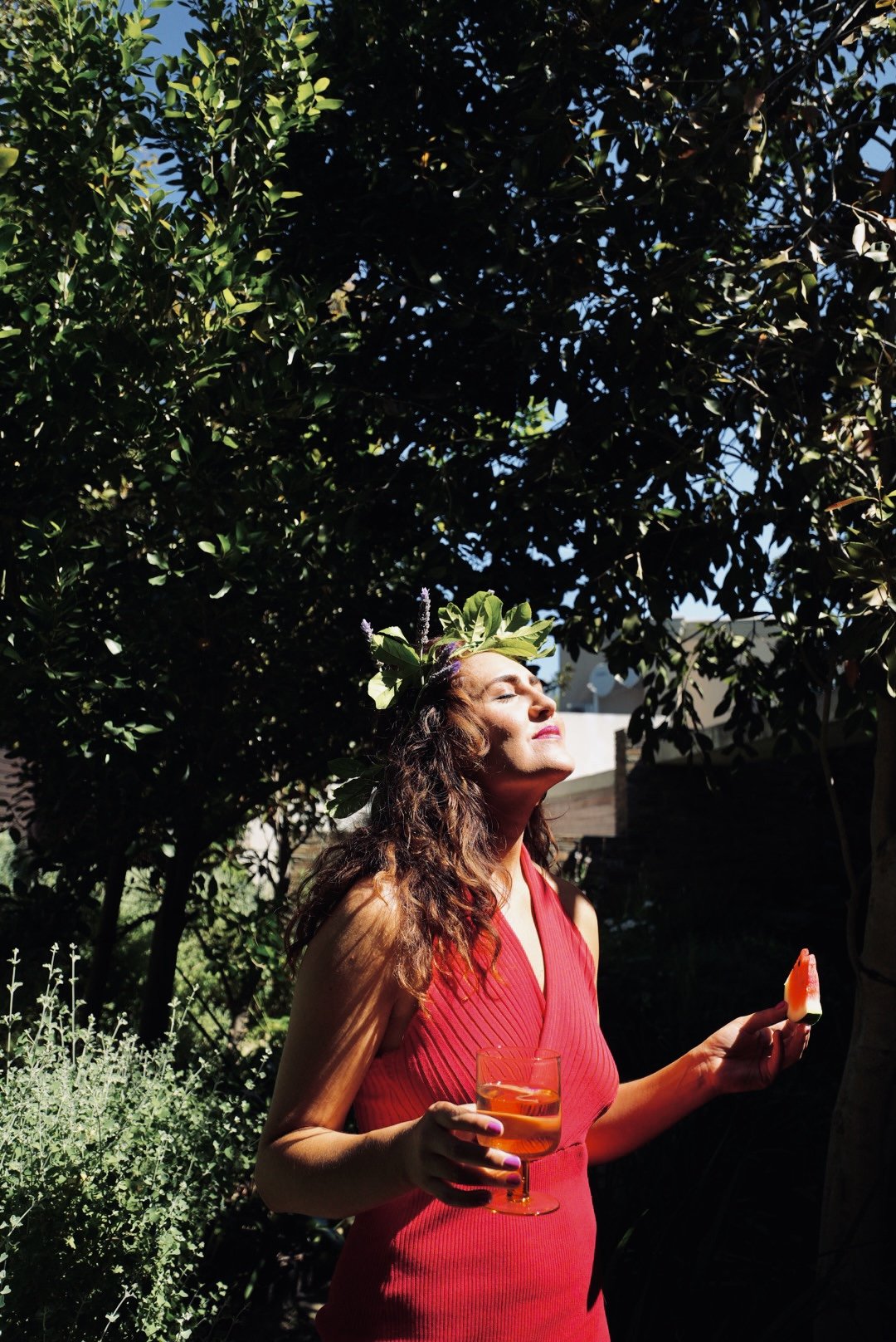 A woman wearing a red dress and a leafy headband stands outdoors, holding a glass of orange drink and a small piece of watermelon, with her eyes closed and face tilted toward the sun.